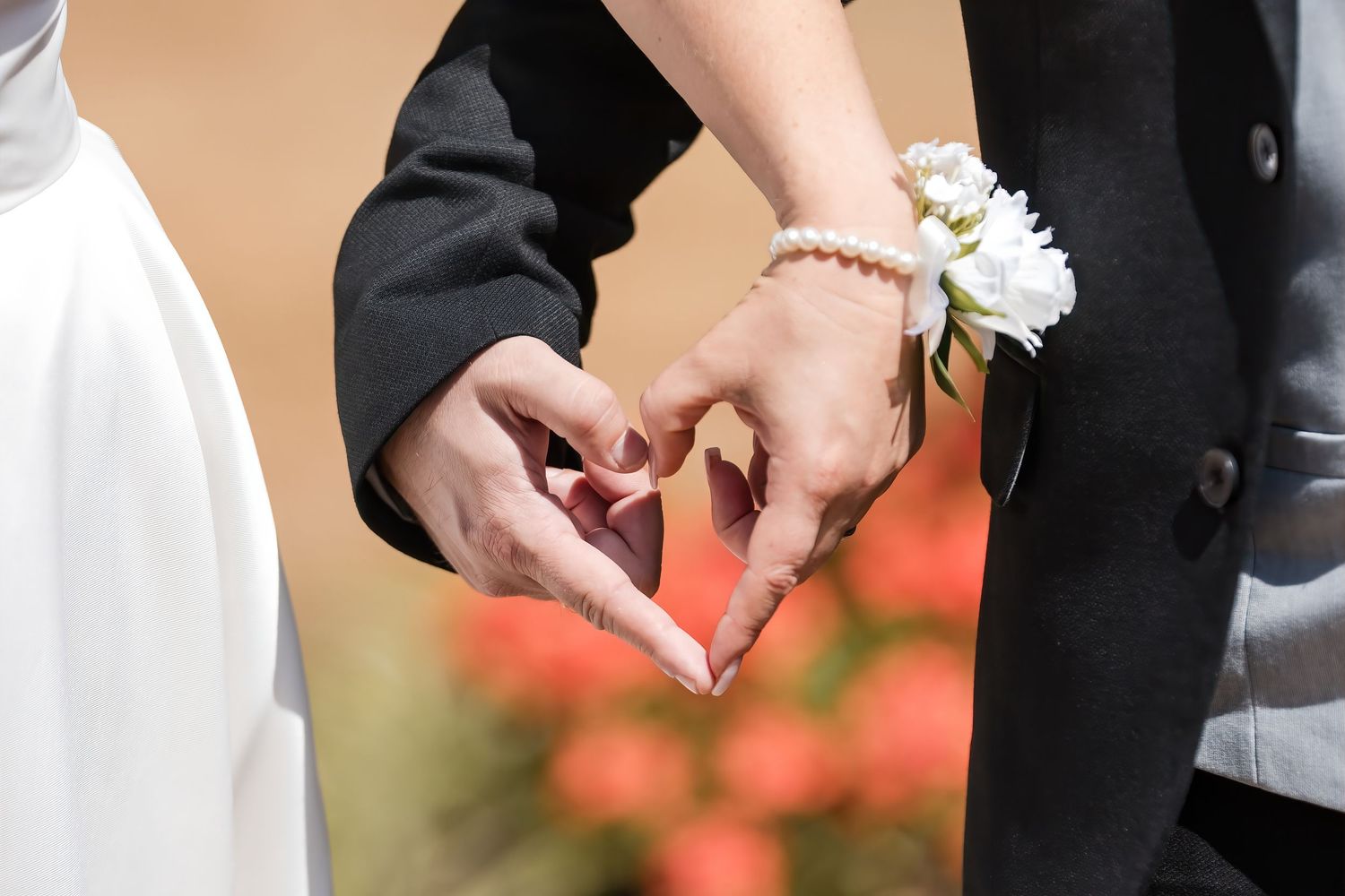 Close-up of wedding couple forming heart shape with their hands, bride wearing pearl bracelet and floral corsage.