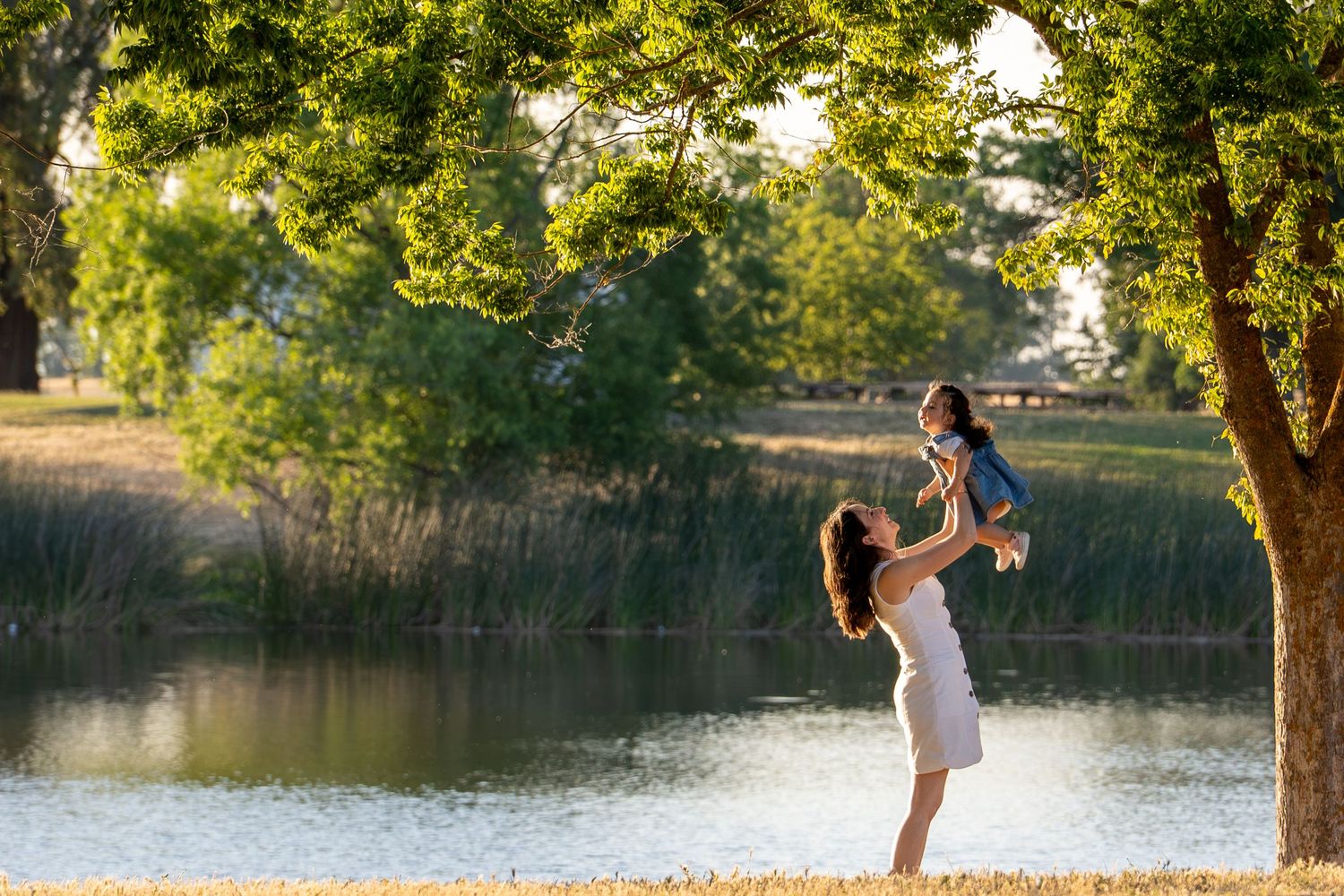 Mom lifting daughter under green tree by the lake at Gibson Ranch, joyful candid family photography during golden hour.