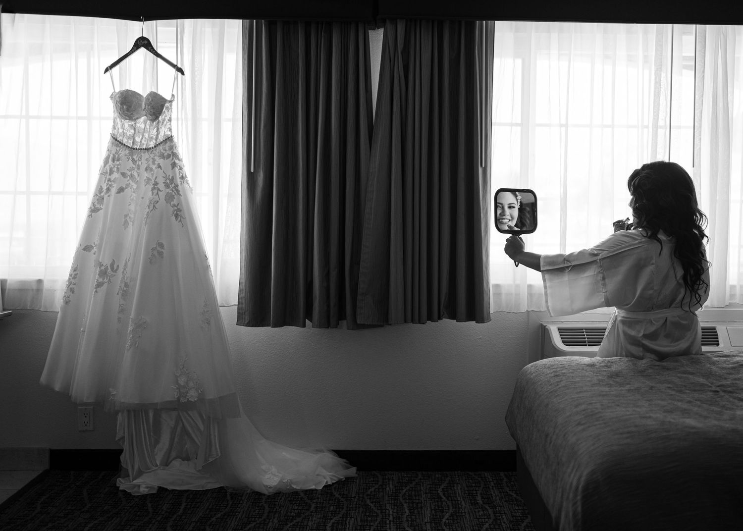 Bride in hotel room holding mirror next to hanging wedding dress, documentary wedding photography by The Padillas.
