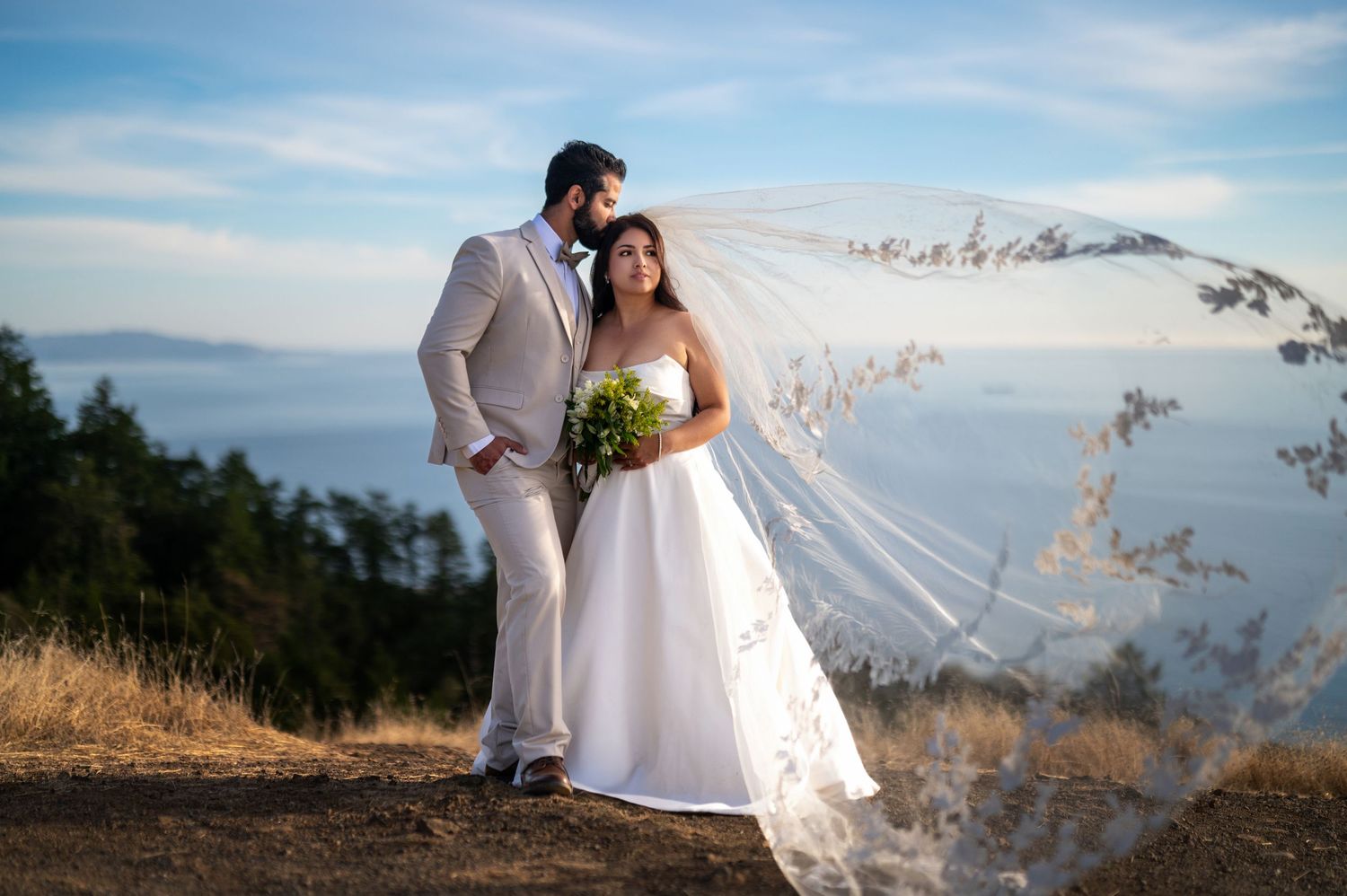 Bride and groom at coastal cliff with flowing veil, elegant destination wedding photography with ocean backdrop