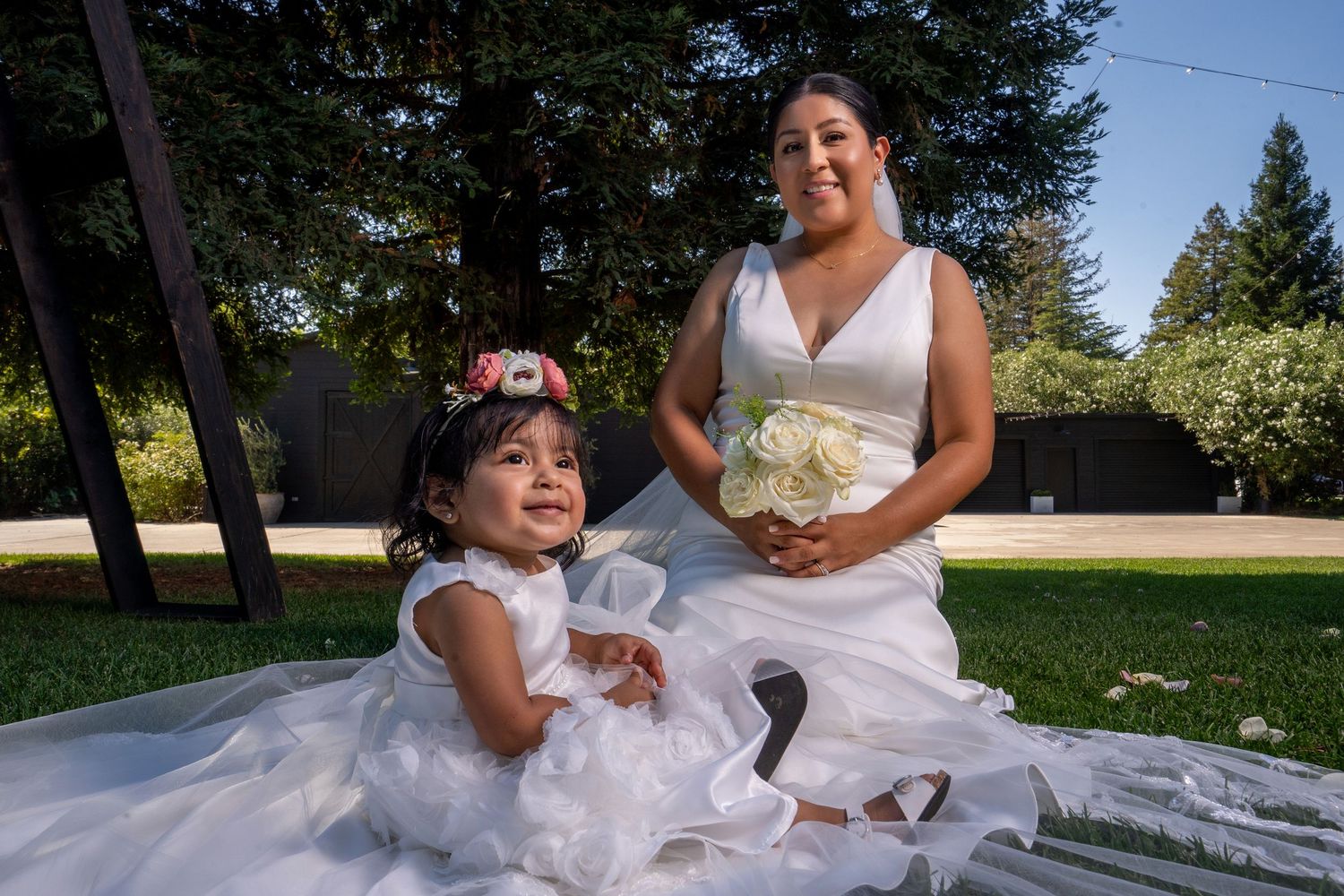 Bride and flower girl sitting together in wedding dress on grass at The Ranch at Lone Oak, photographed by The Padillas.