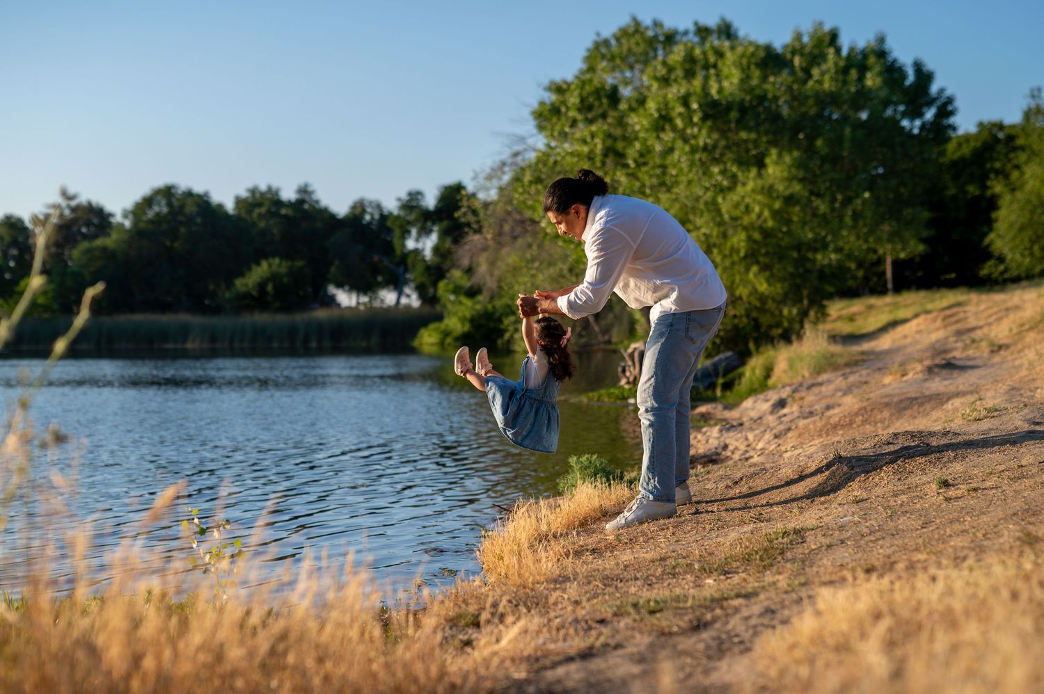 Dad swings daughter by the lake at Gibson Ranch during golden hour, playful candid family photography.