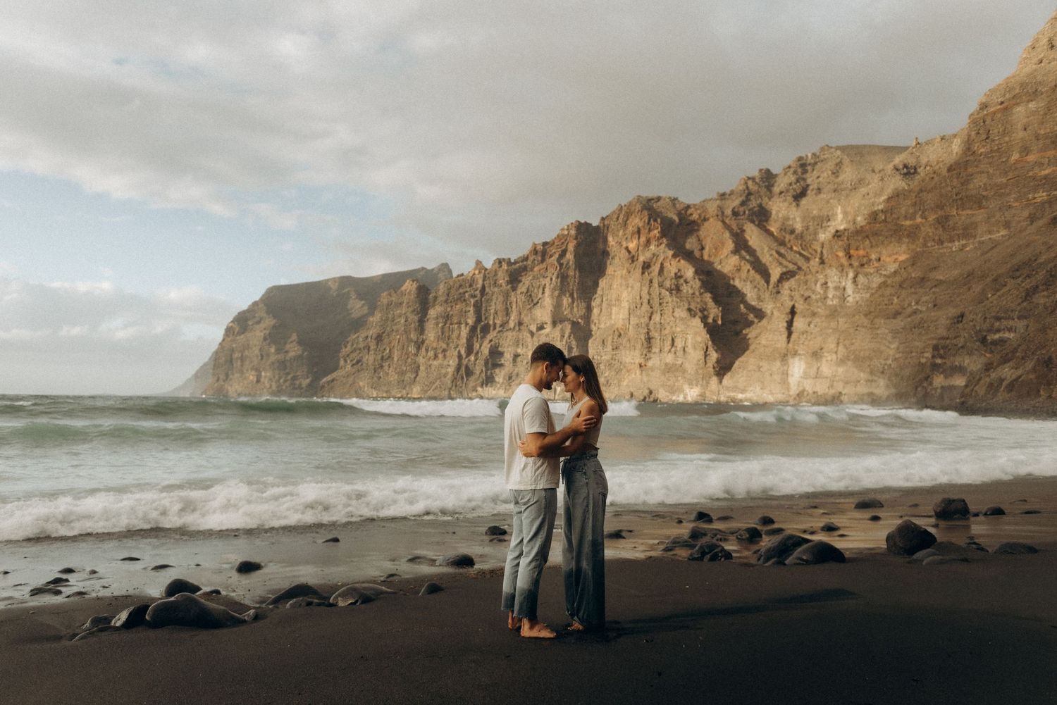Young couple in romantic embrace on Playa de Los Guíos with dramatic Los Gigantes cliffs, Tenerife