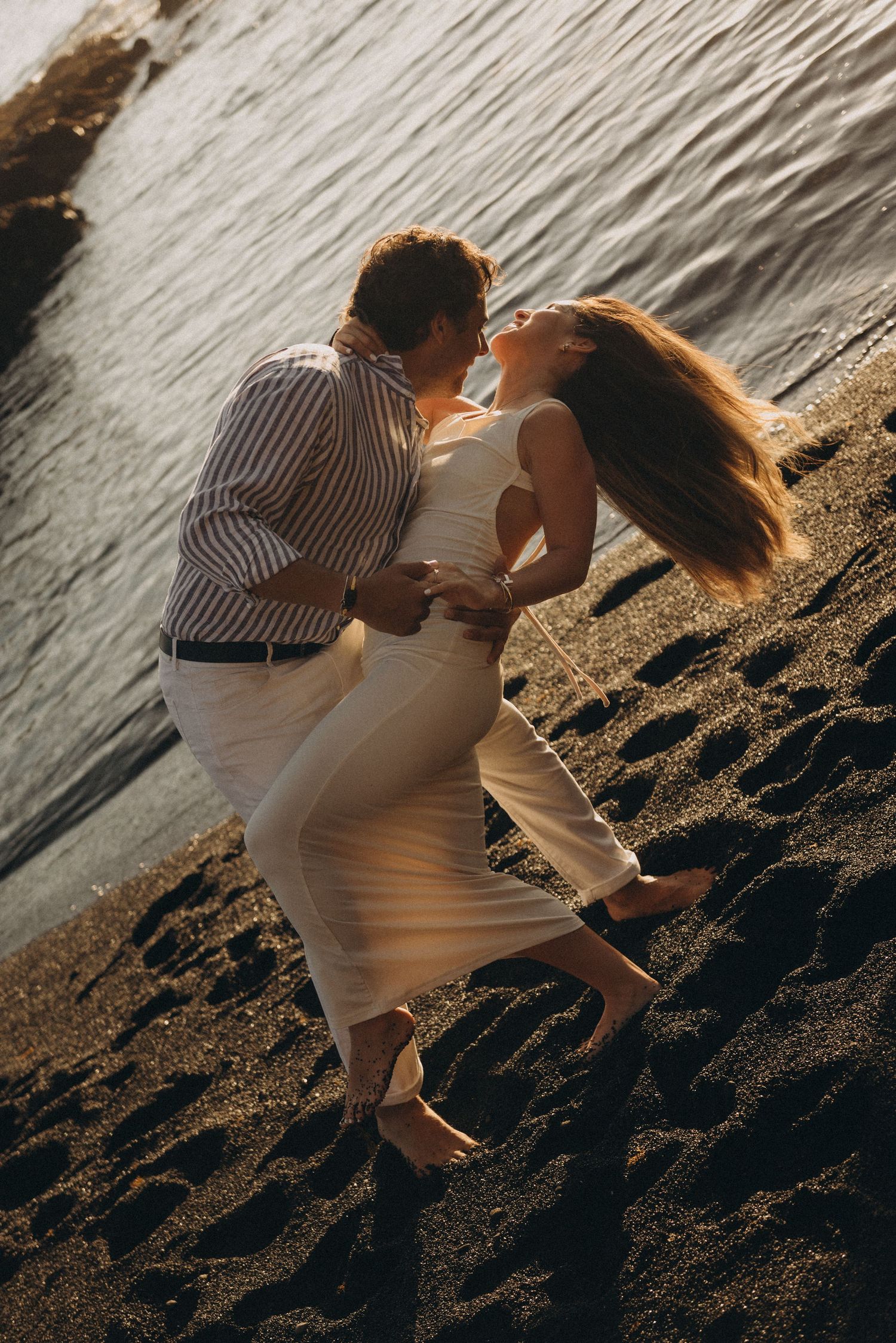 Romantic couple embracing and kissing on a dark sandy beach at golden hour by the ocean.