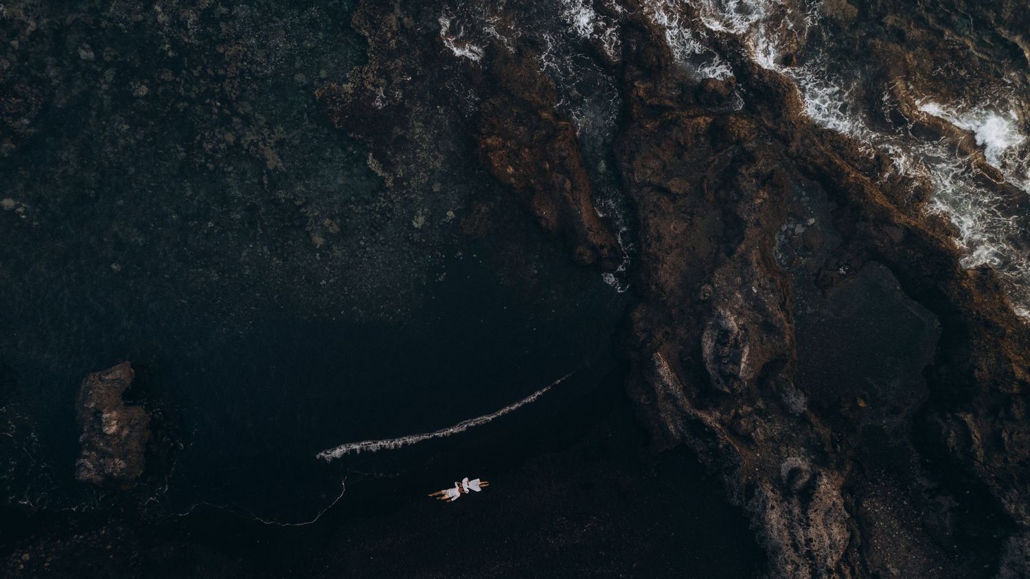 Aerial view of a small boat navigating dark rocky coastal waters from above.