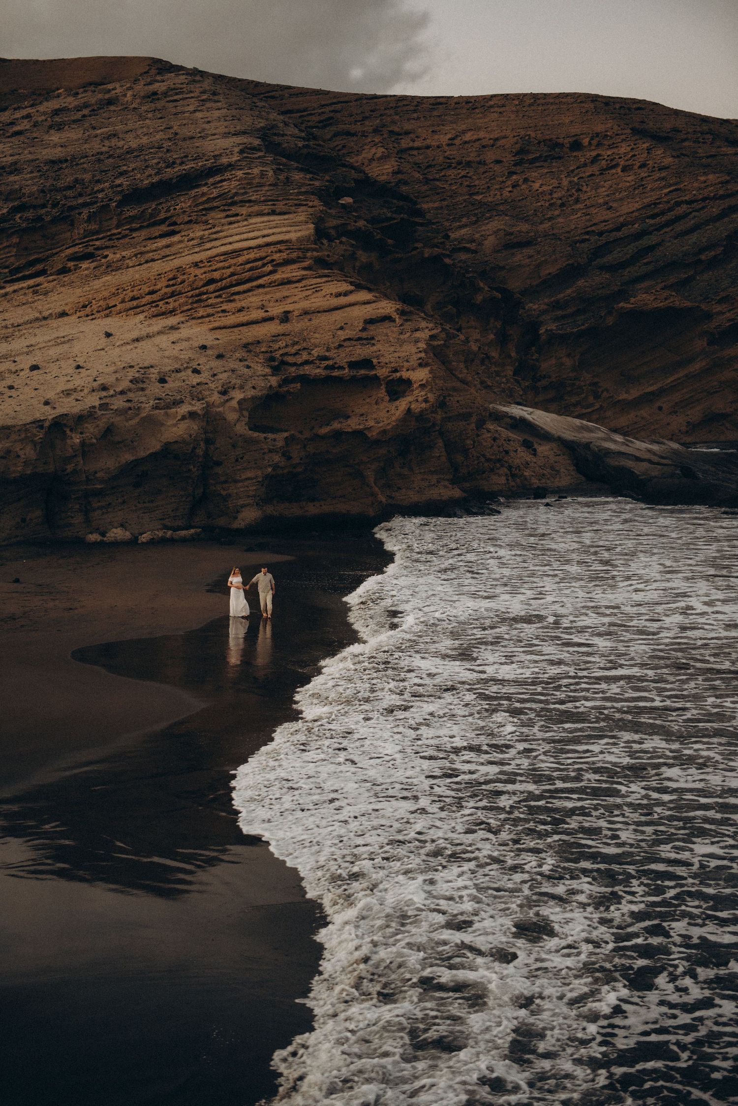 Couple in wedding attire standing on a black sand beach with dramatic ocean waves and rugged volcanic cliffs.