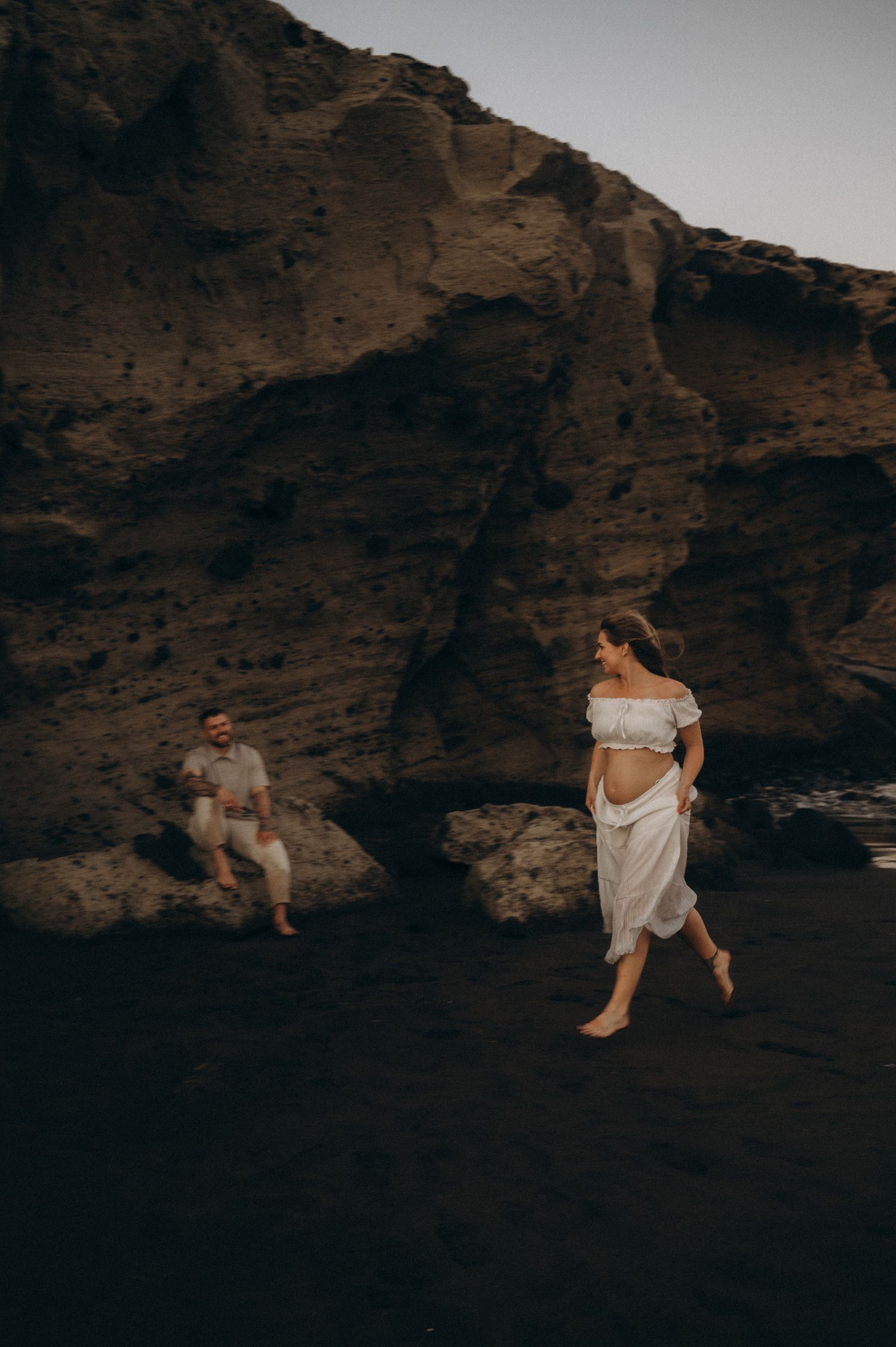 A pregnant woman in a white dress walks on a black sand beach near rocky cliffs as a man sits watching her.