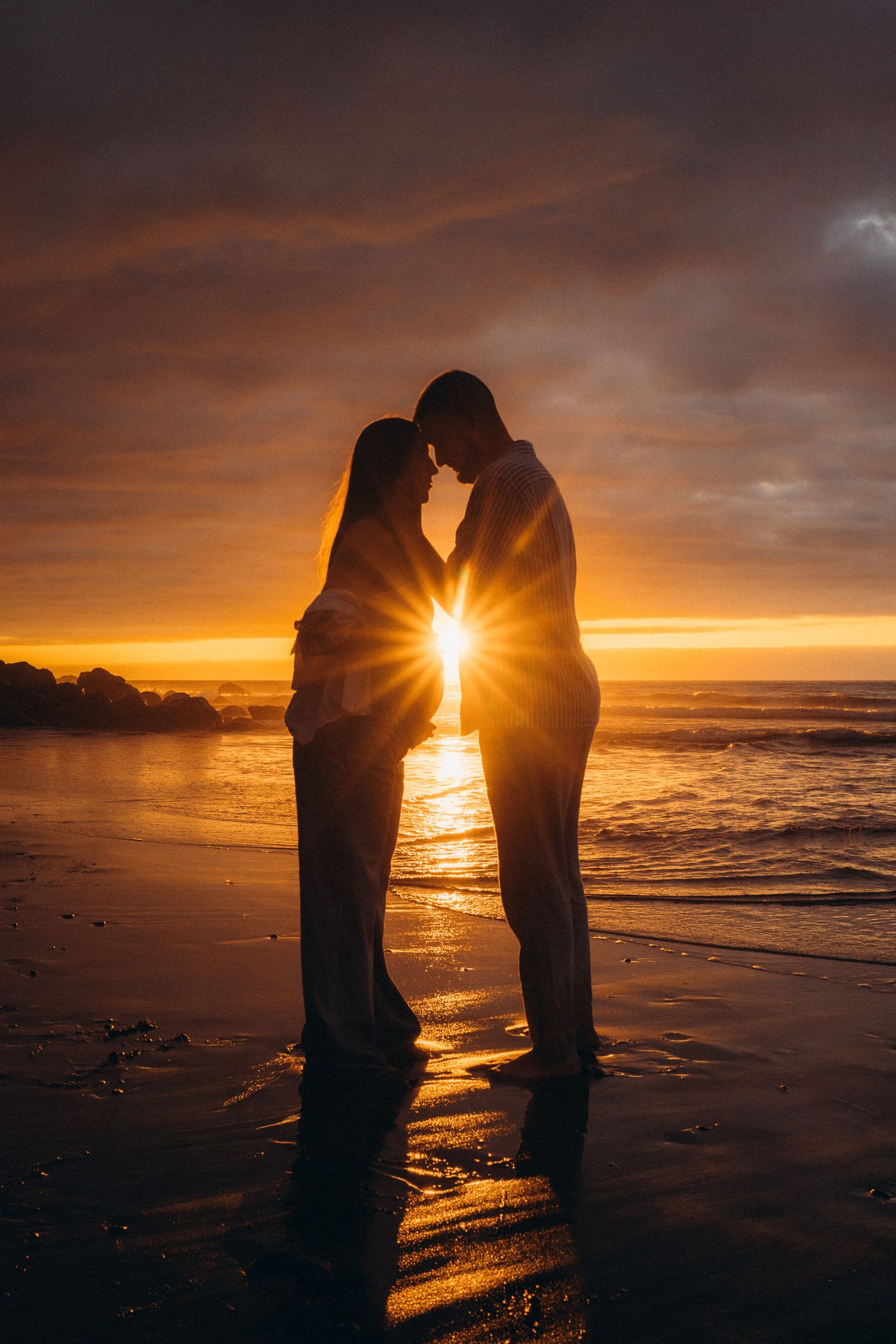 Romantic couple silhouetted against a stunning golden sunset on a beach, with sun rays glowing between them.