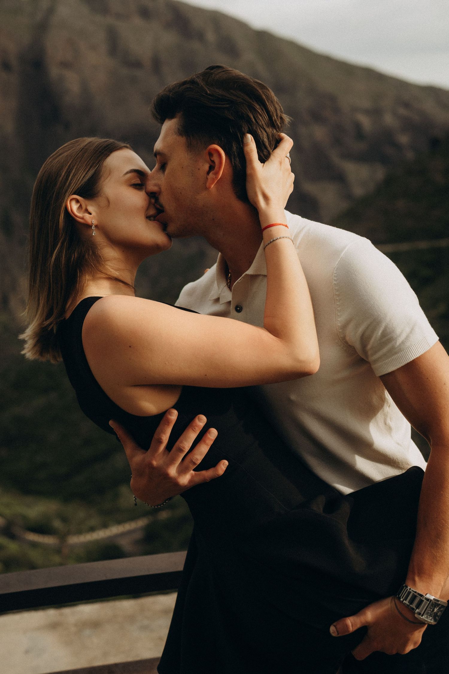 A romantic couple shares a passionate kiss on a scenic mountain balcony, dressed in black and white outfits.
