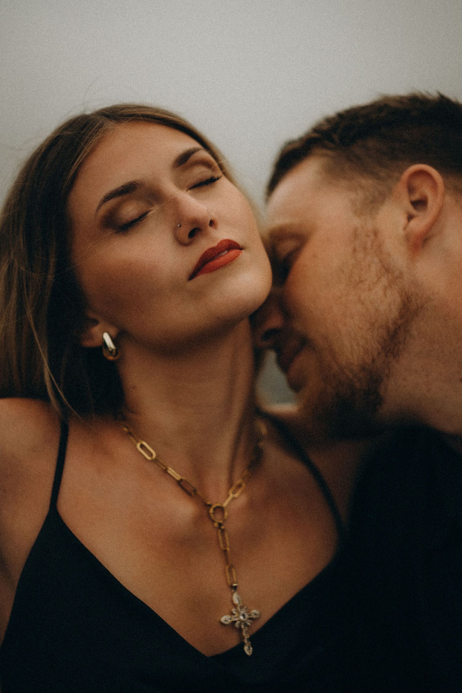 A couple in an intimate moment, woman wearing a gold cross necklace and hoop earrings with red lips, eyes closed.