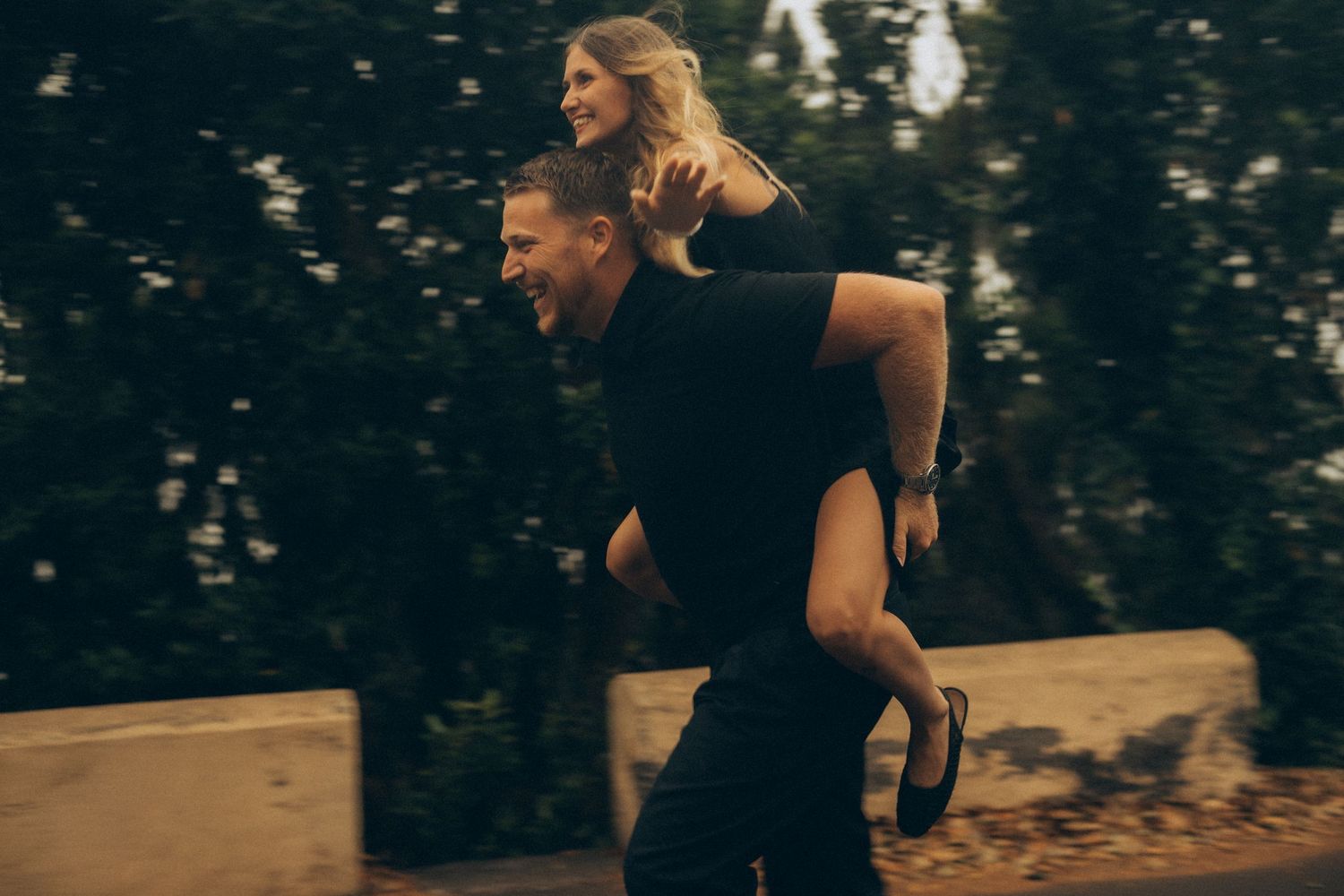 A man in black gives a woman a piggyback ride, both laughing outdoors near lush green trees.