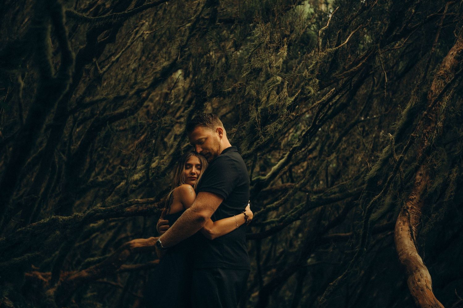 Couple embracing romantically in front of dramatic dark twisted tree roots in a lush forest setting.