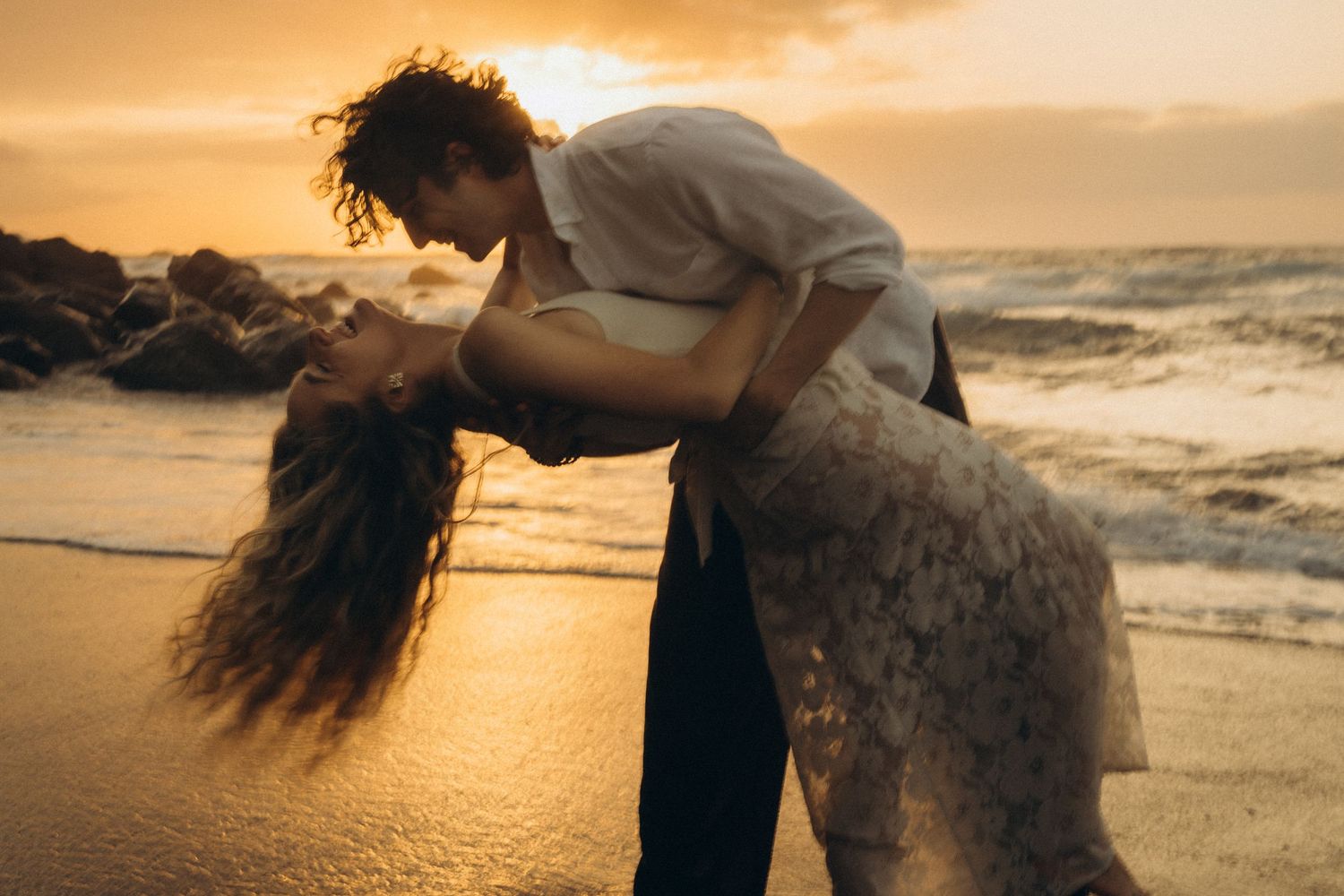 Couple sharing a romantic dip kiss on a sandy beach at golden sunset with ocean waves in the background.
