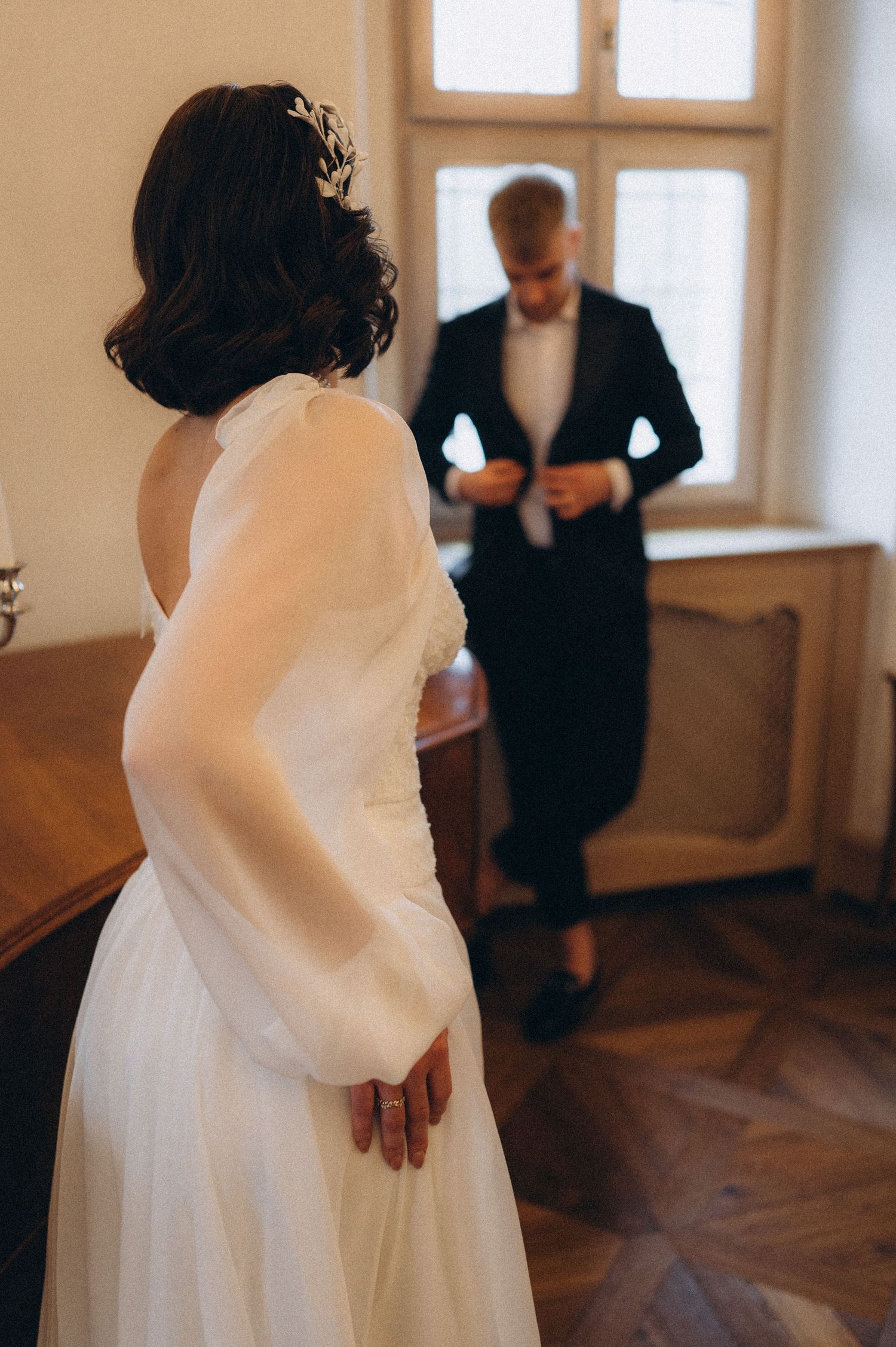 Bride admiring the groom from a distance as he buttons his suit jacket during their wedding photoshoot.