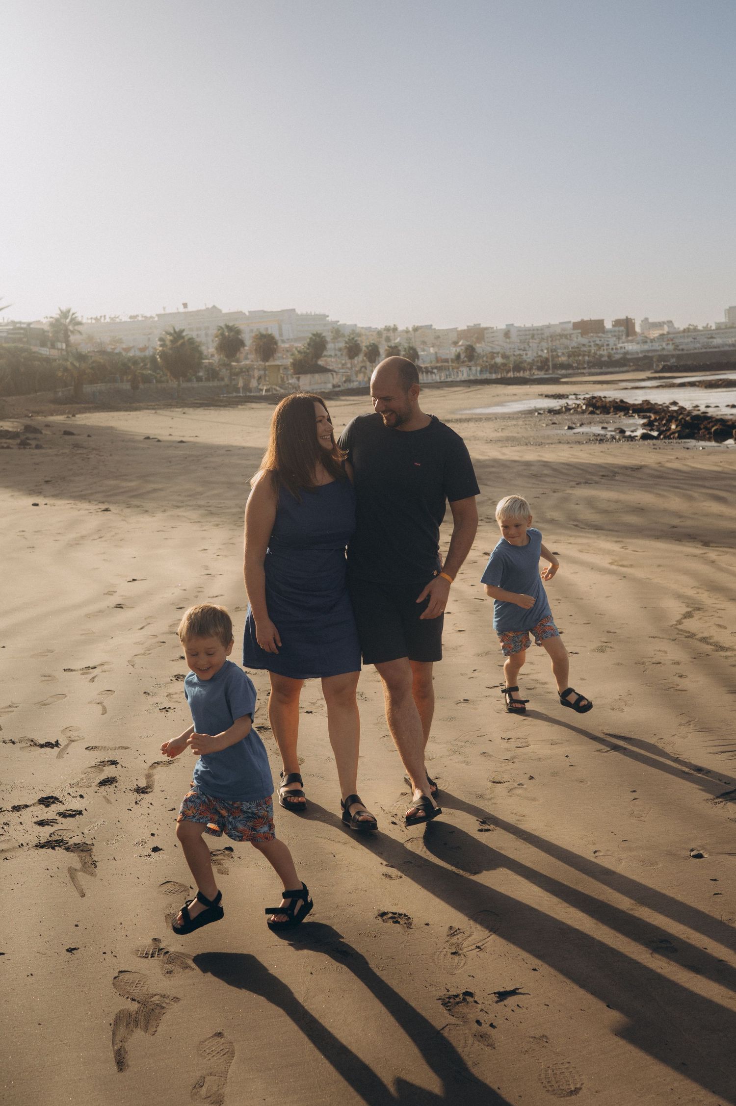 Parents walking on warm golden sand at sunrise while their children run around them at Playa Fañabé, storytelling family photography