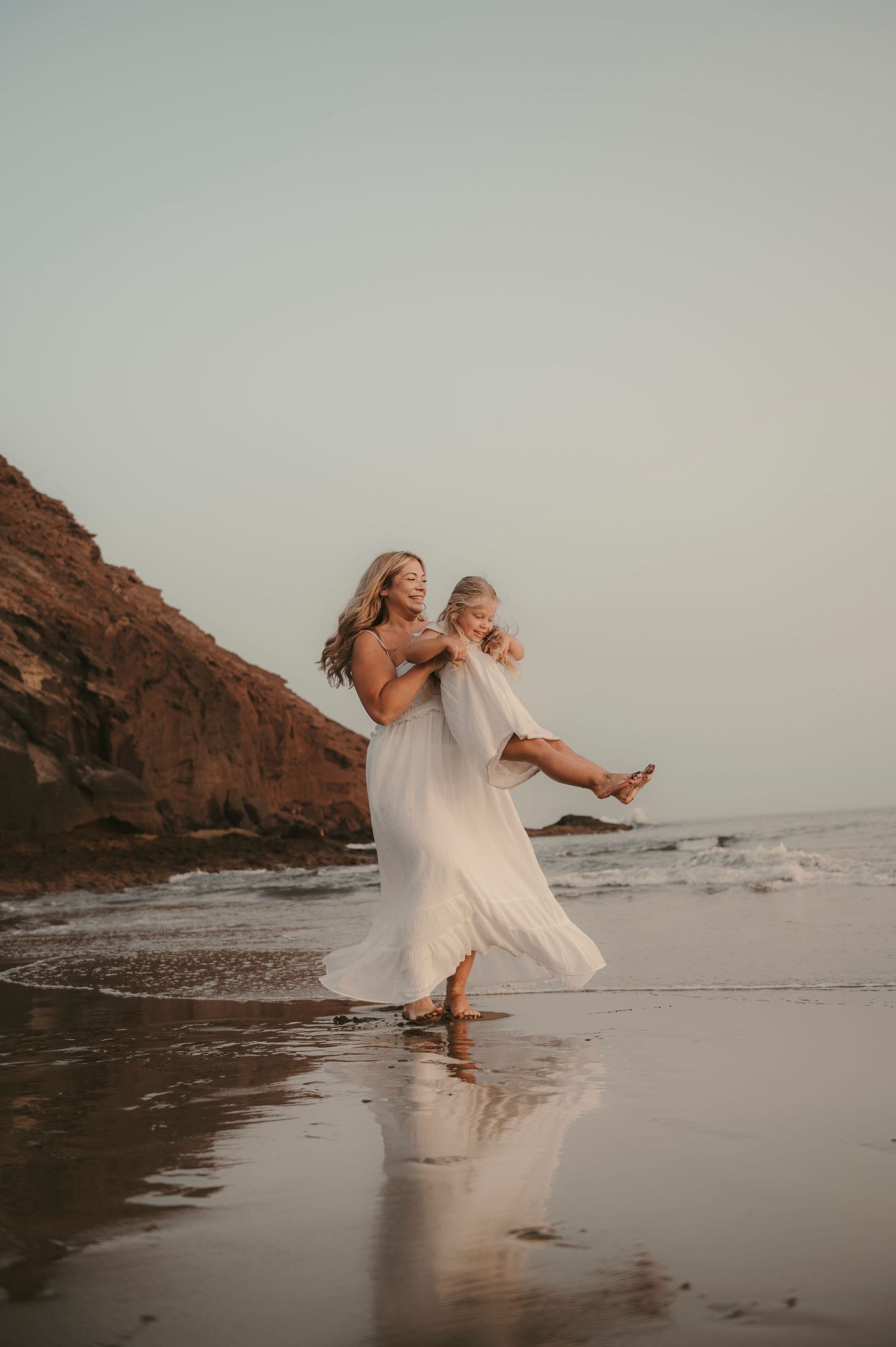 Mother holding her daughter while twirling together with reflections in the sand, ocean and Montaña Roja