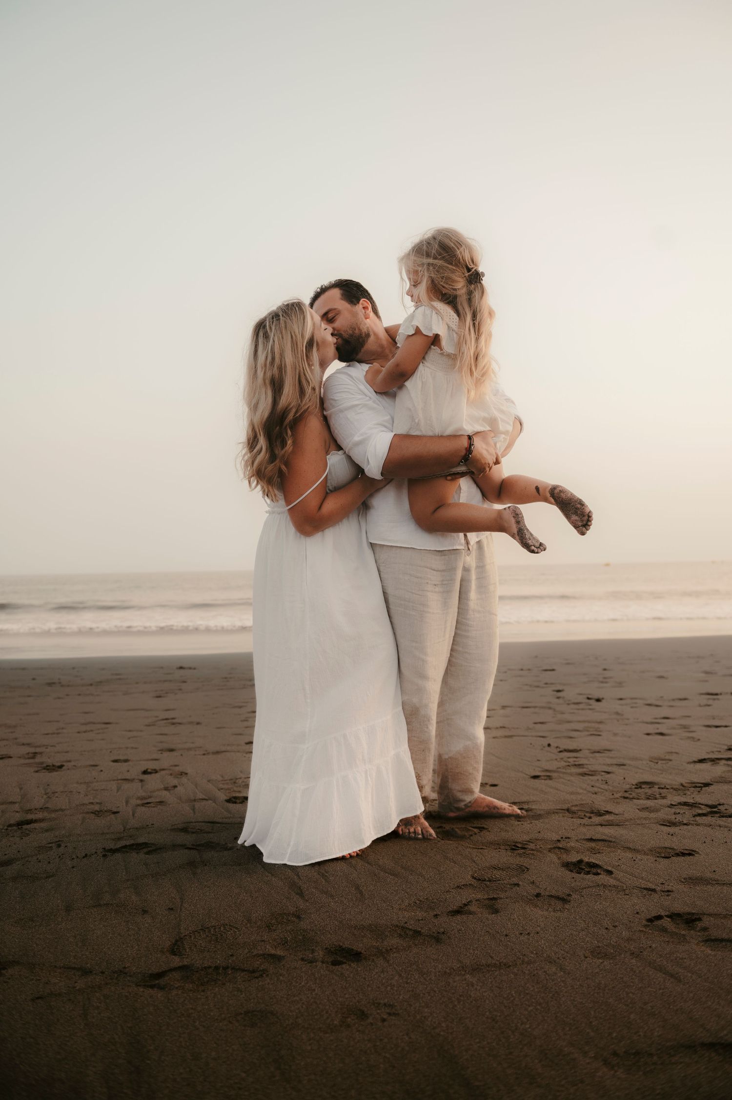 Tender family moment with mother and father kissing as their little girl is in dad’s arms on Playa La Tejita beach at golden hour