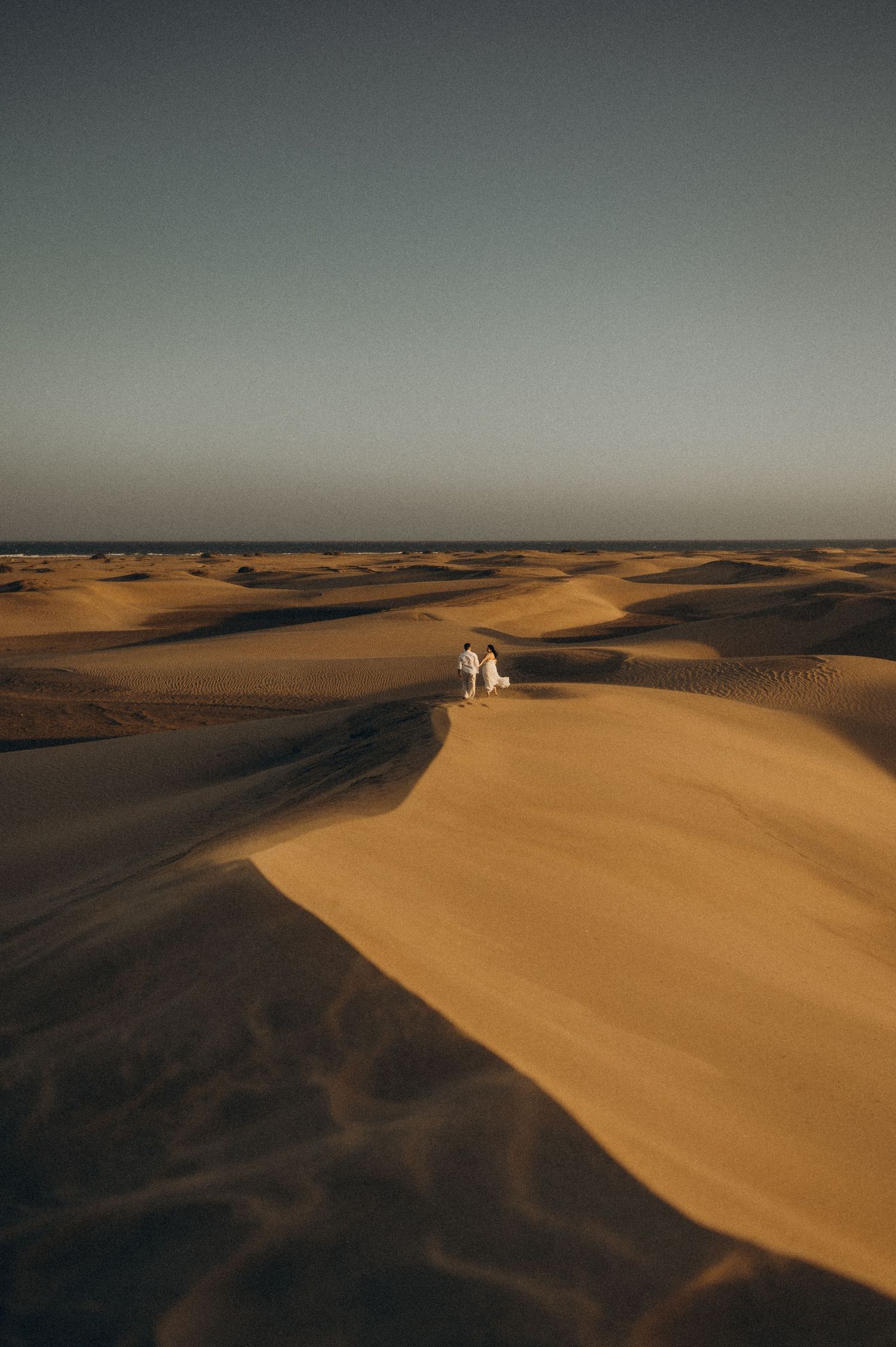 Couple walk among vast golden sand dunes under a clear sky in a serene desert landscape at golden hour. Captured in Tenerife.