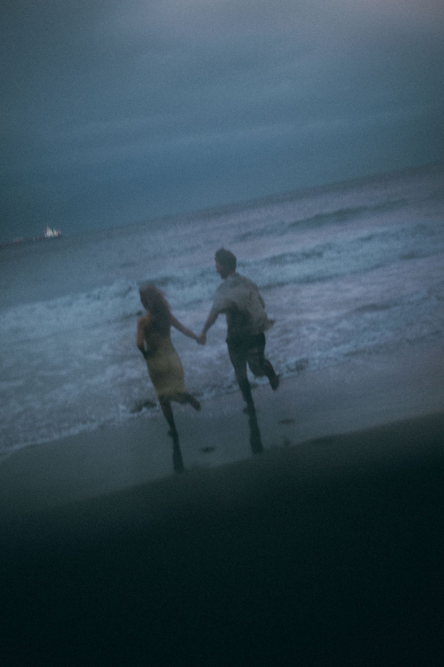Couple holding hands running on a dark beach at dusk with ocean waves in the background.