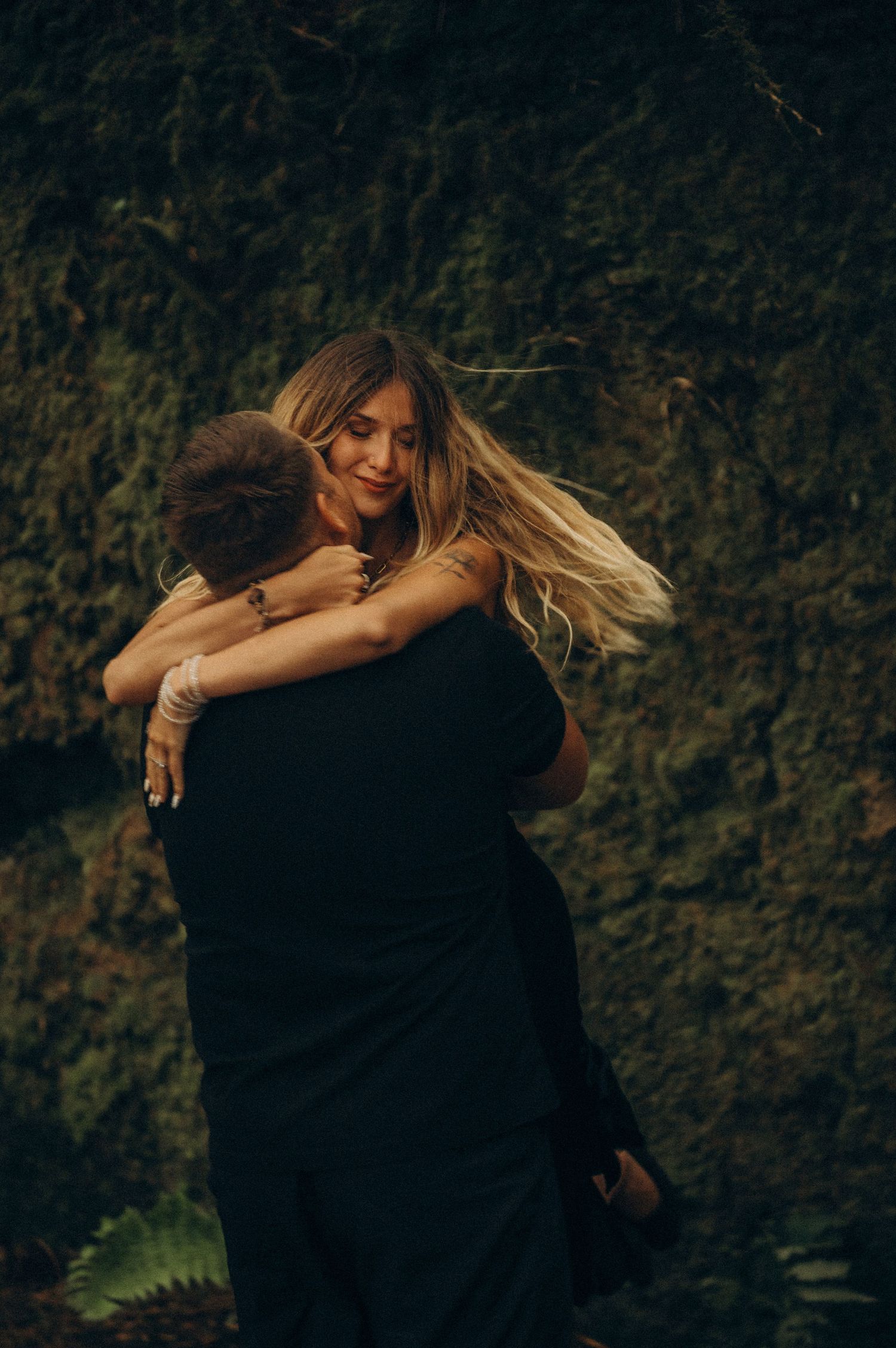 A couple embraces romantically in front of a lush green ivy wall, the woman smiling with flowing blonde hair.