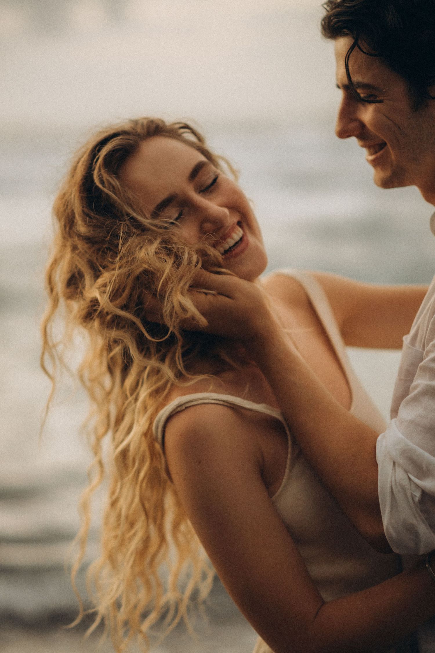 A smiling couple shares a tender moment by the ocean, with the woman laughing as the man gently touches her face.