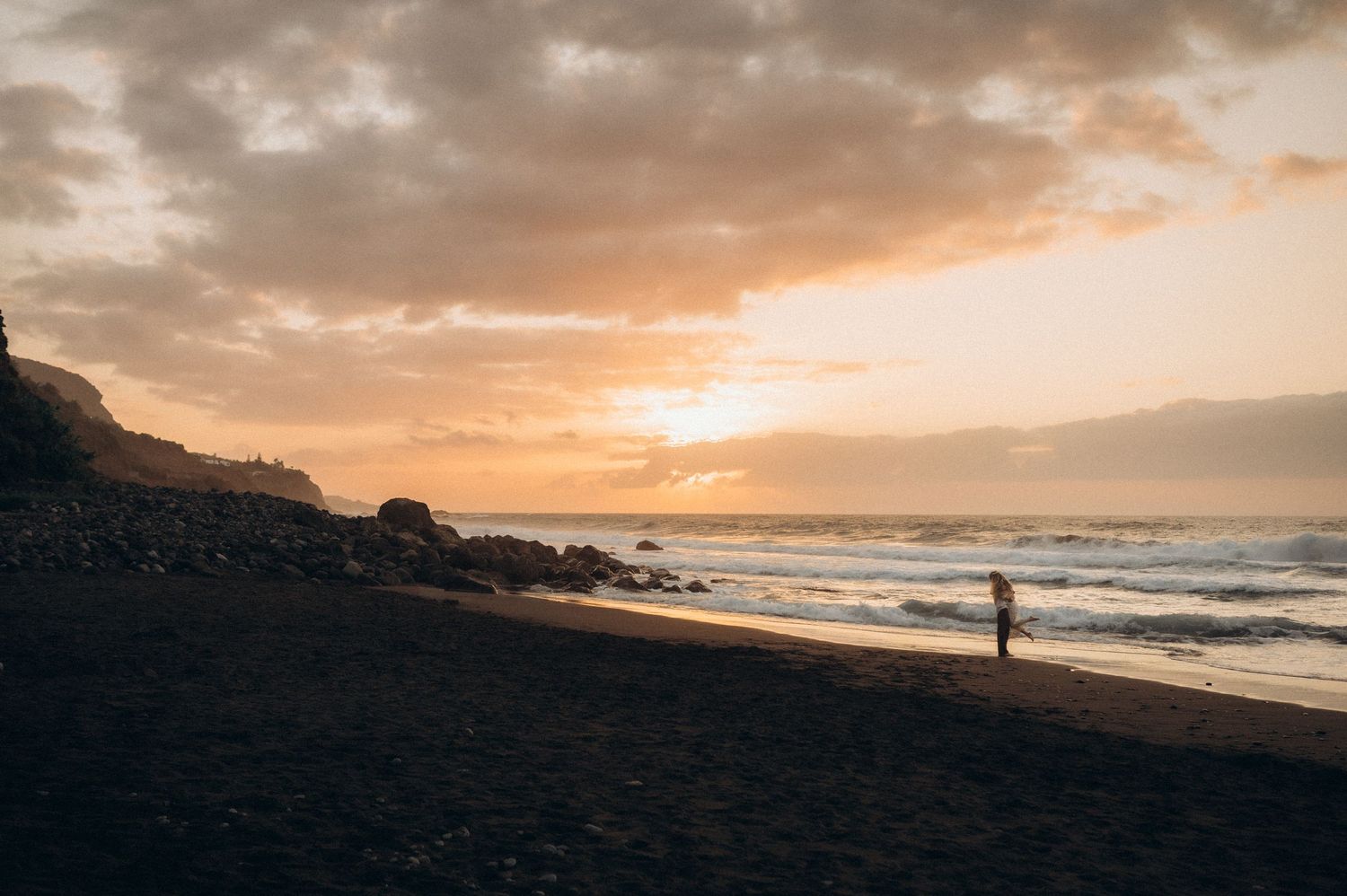 A lone figure stands on a dark sandy beach at sunset, with golden clouds over the ocean and rocky cliffs nearby.