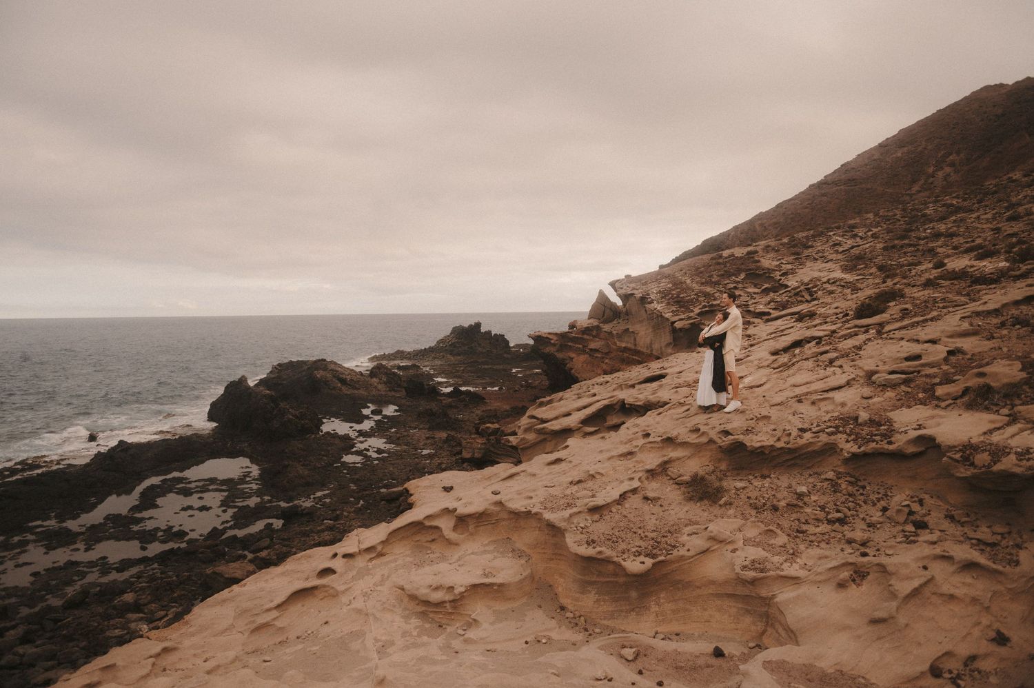 Cinematic shot of a couple standing on the rocks under Montaña Roja, enjoying the breathtaking ocean and cliff views in the background, Tene