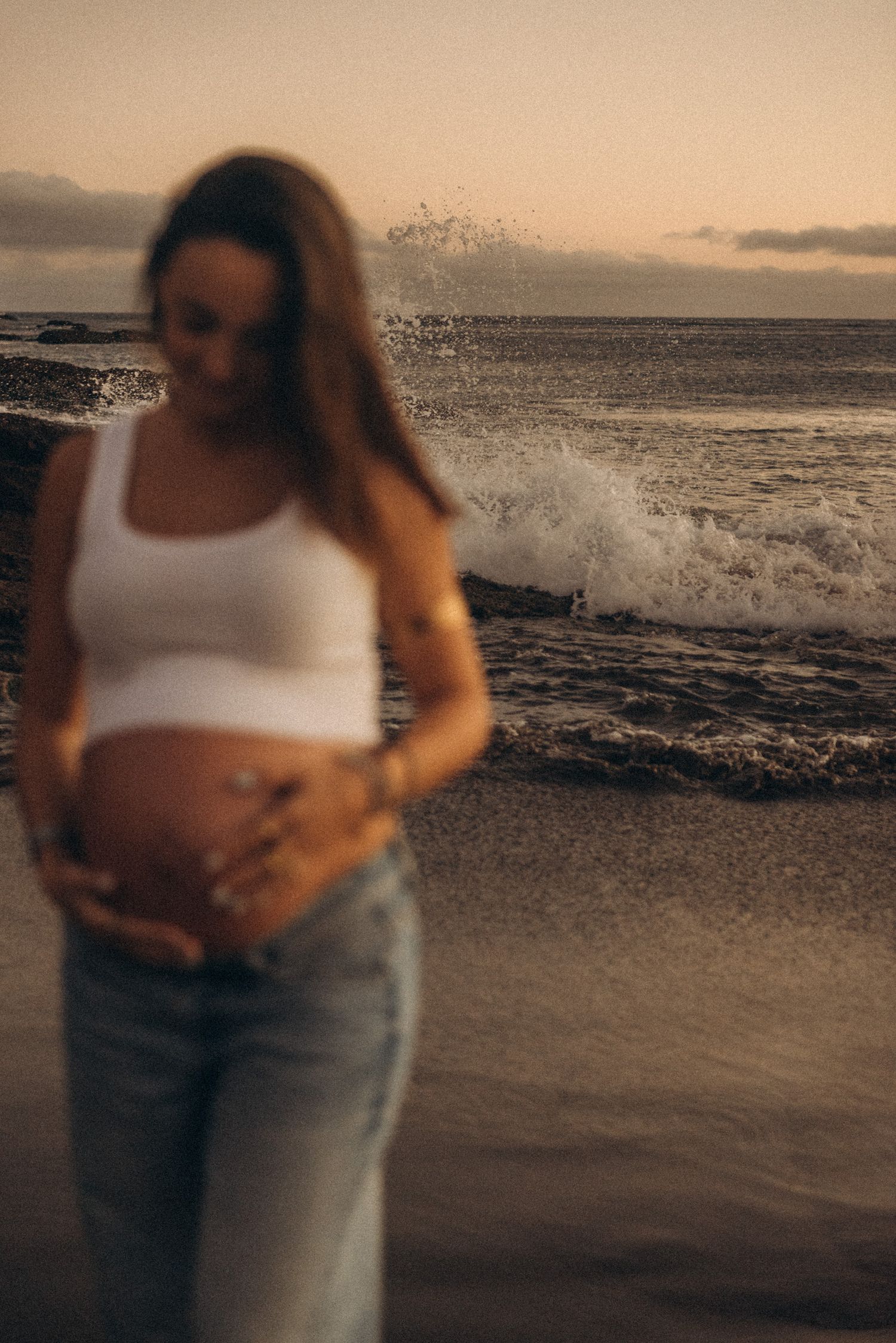 Pregnant woman in white crop top and jeans cradling her baby bump on a beach at sunset with waves crashing.