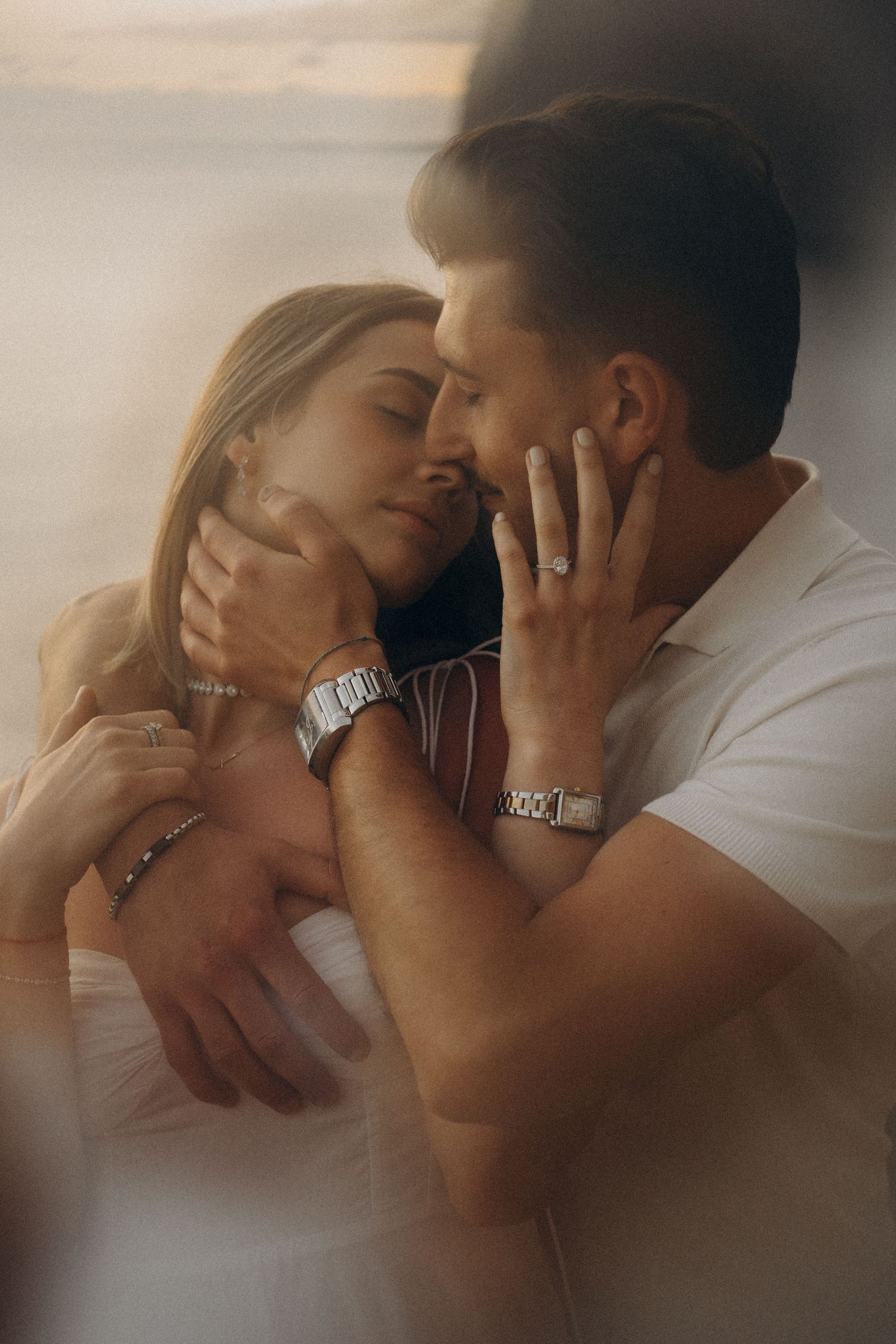 A romantic couple embraces tenderly, the woman wearing an engagement ring, both adorned with silver jewelry in warm golden light.