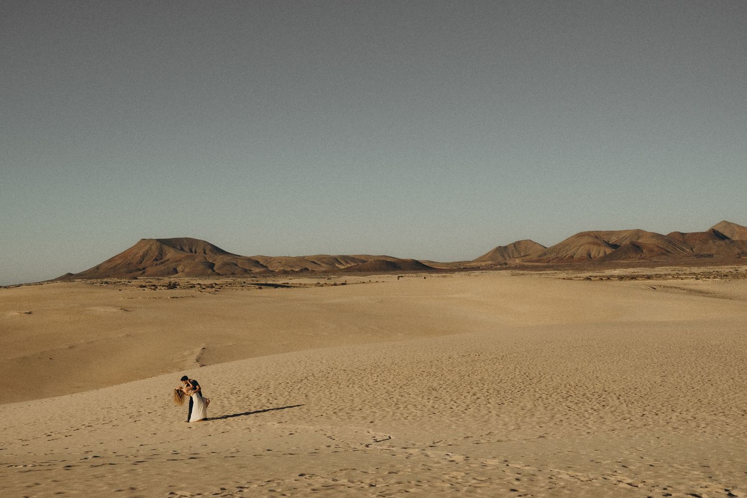 A lone figure walks across vast golden sand dunes with rocky mountains in the background under a clear desert sky.