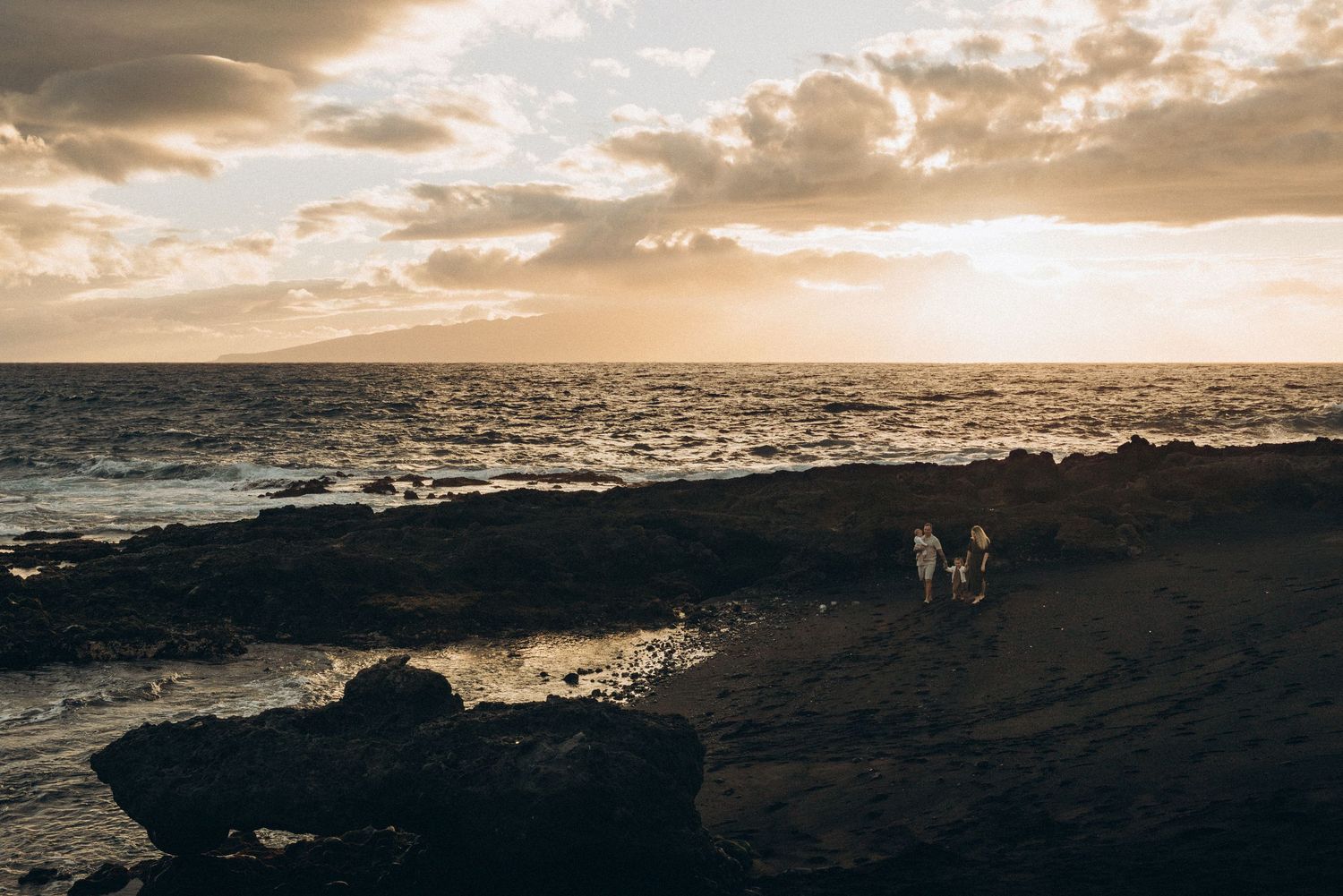 Two people walk along a rocky coastline at golden sunset with dramatic clouds and ocean waves in the background.
