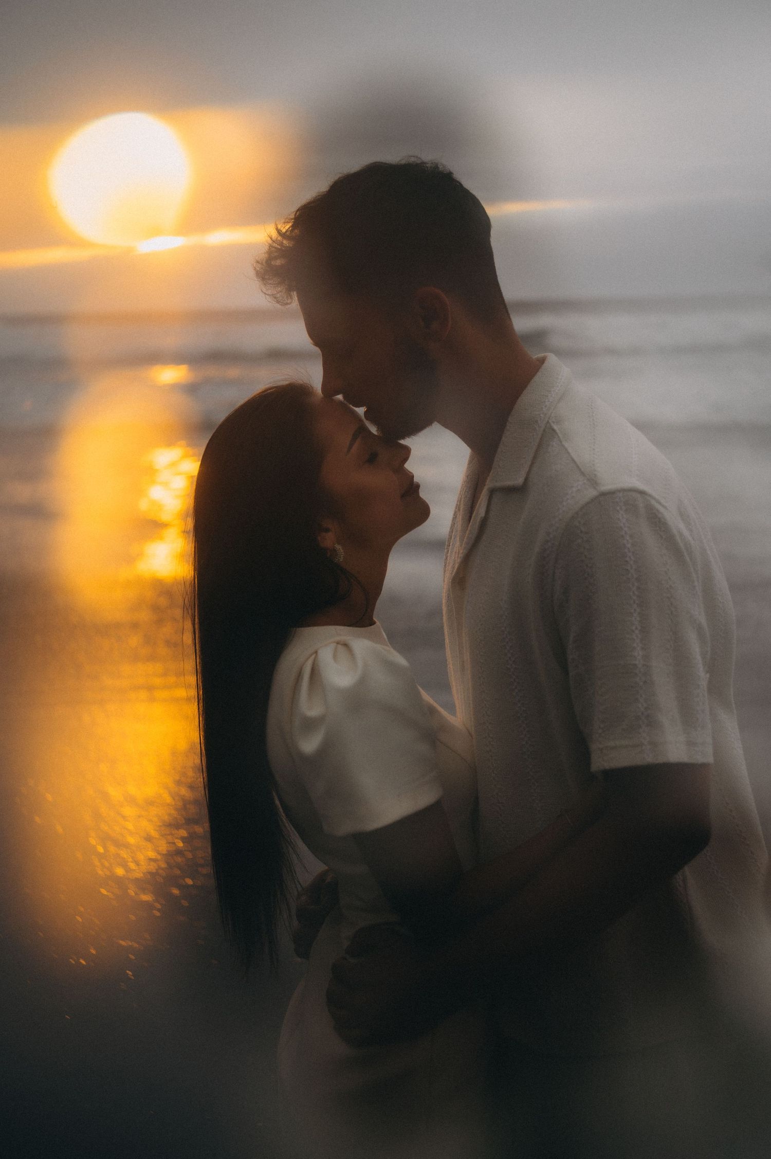 Couple sharing a romantic almost-kiss at sunset on the beach, golden light reflecting on the ocean waves behind them.