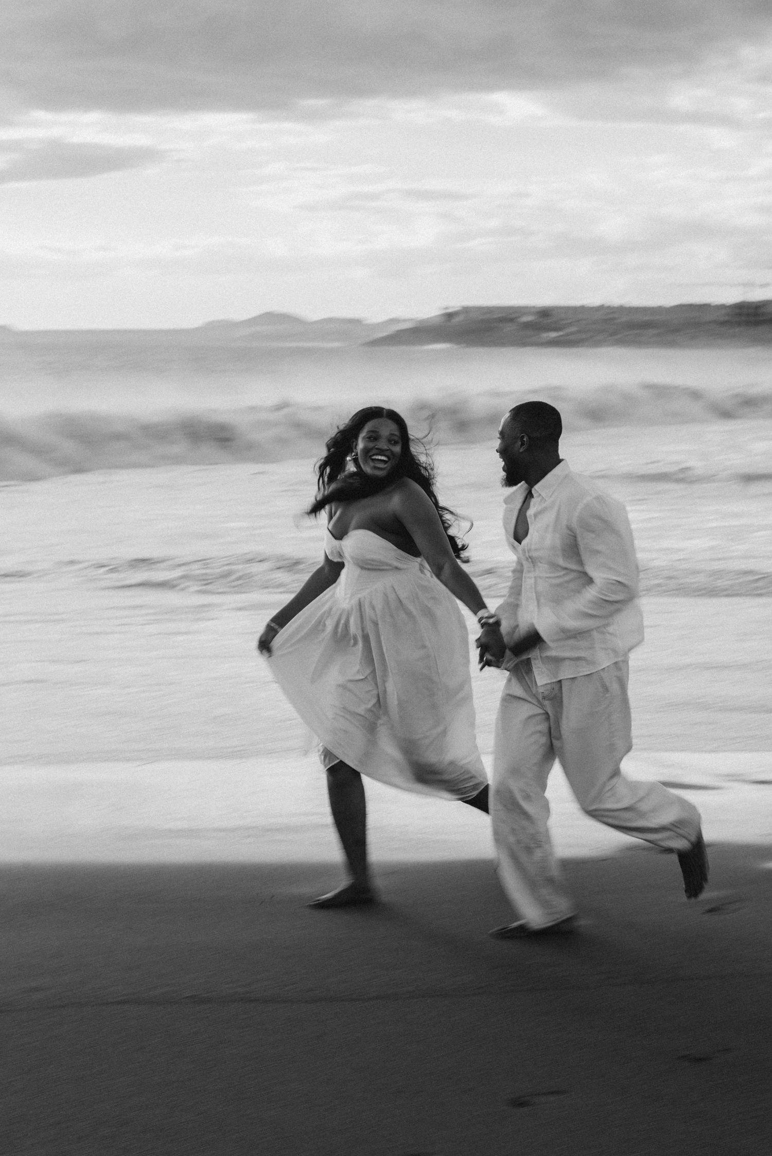 Two people in flowing white dresses running and dancing on misty Tenerife beach at dusk creating dreamy silhouettes during photoshoot