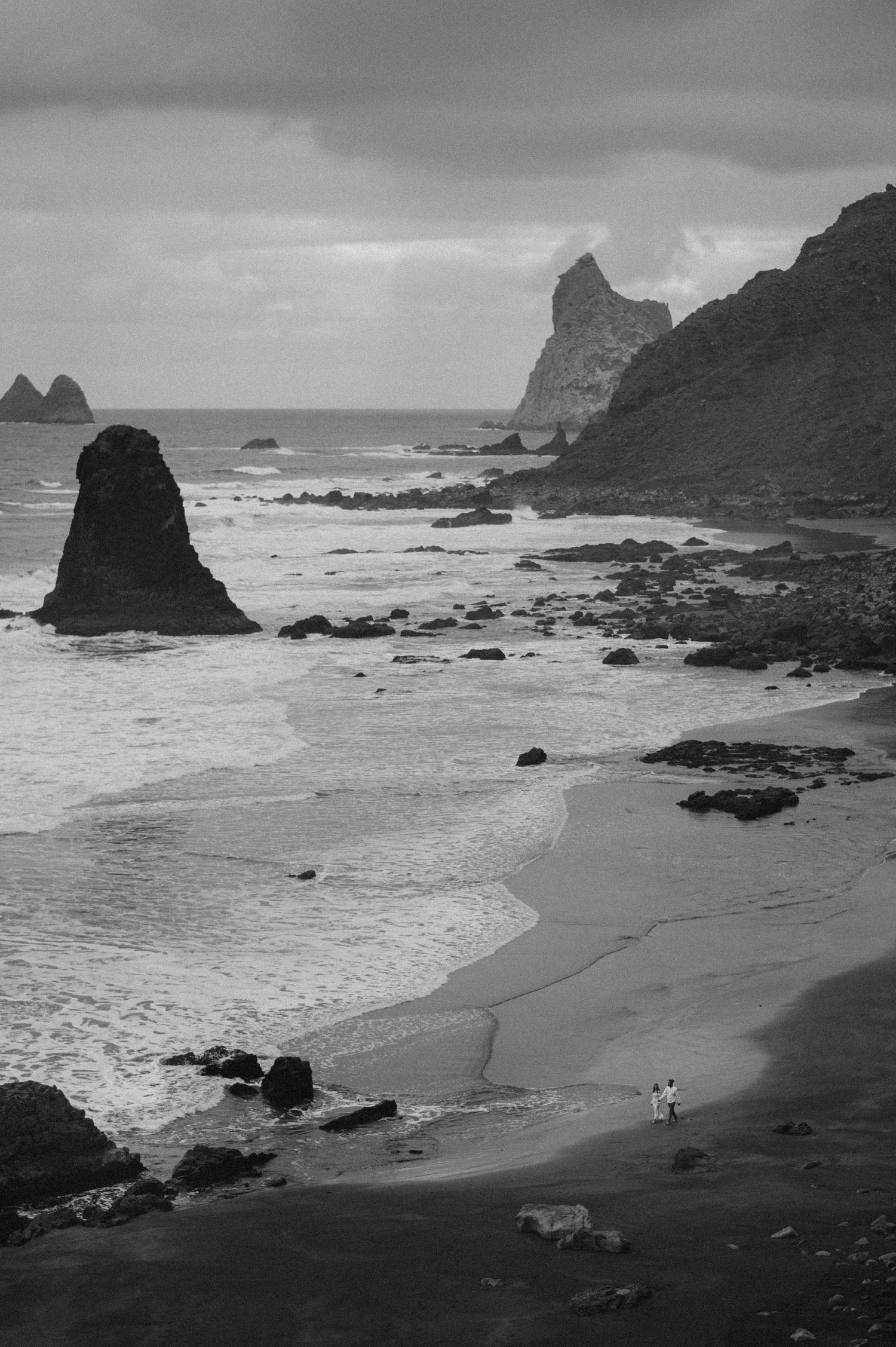 Dramatic black and white panorama of a rugged coastal beach with crashing waves and towering rock formations along the shoreline.