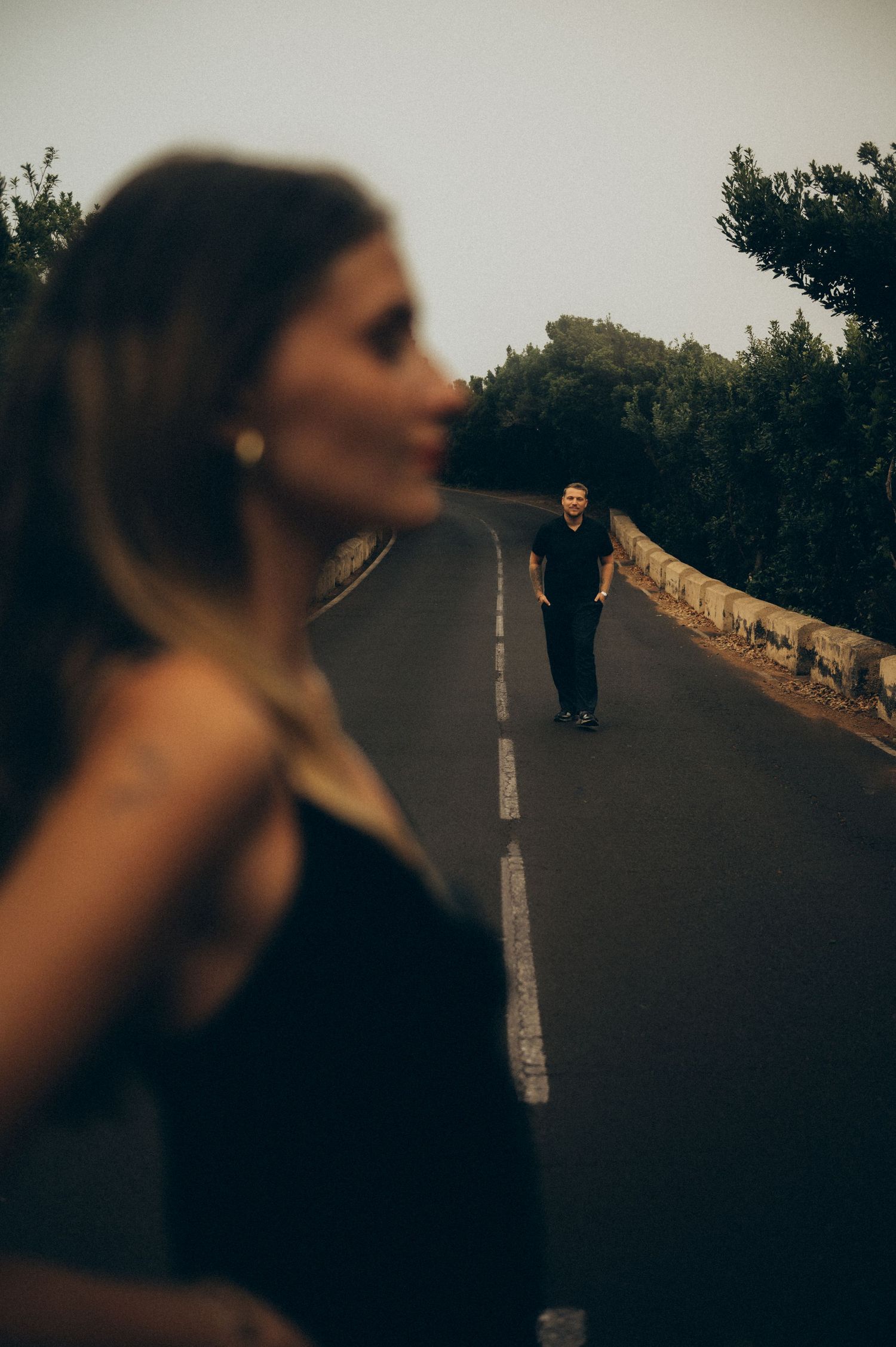 A blurred woman in black stands in the foreground as a man walks down a tree-lined road in moody light.