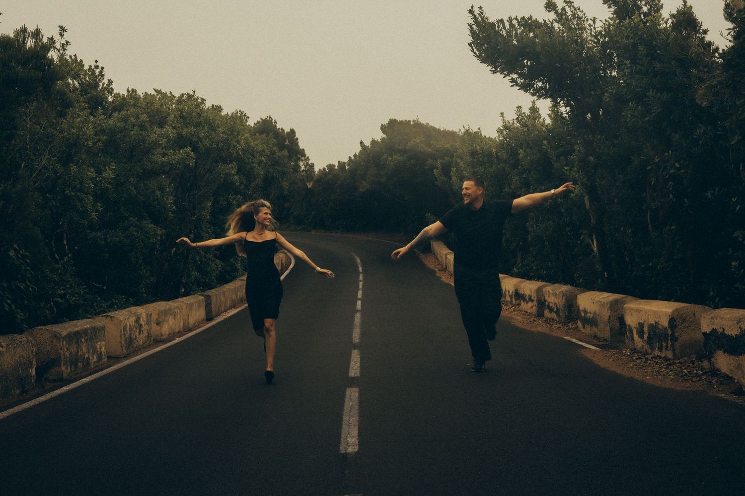 Couple in black outfits dancing on an empty road surrounded by lush green trees in a moody, atmospheric setting.