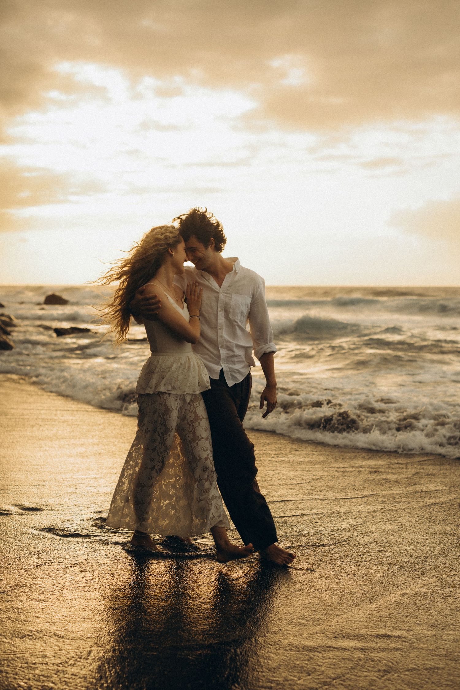Romantic couple embracing barefoot on a sandy beach at golden sunset with ocean waves in the background.