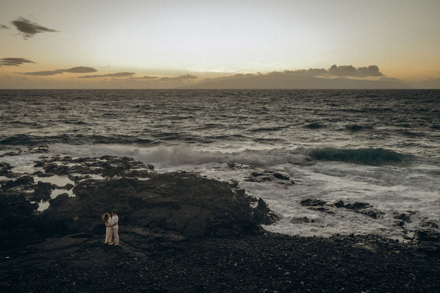 Two figures stand on dark volcanic rocks at sunset overlooking a dramatic ocean coastline in Tenerife
