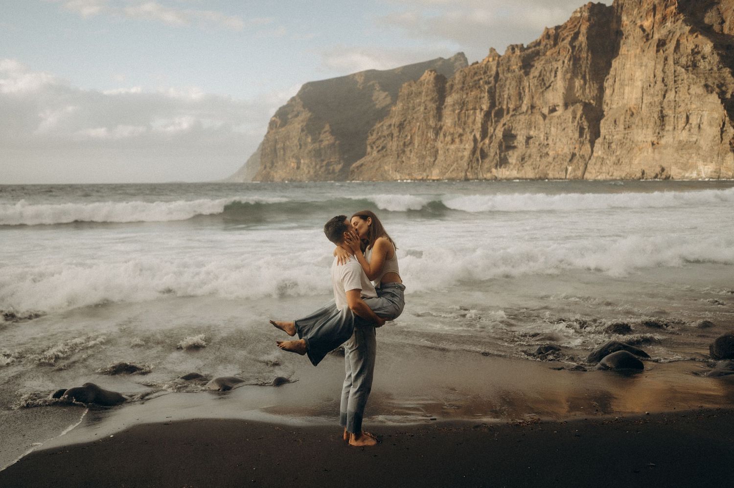 Couple sharing joyful hug as woman jumps into arms on Playa de Los Guíos, Tenerife cliffs