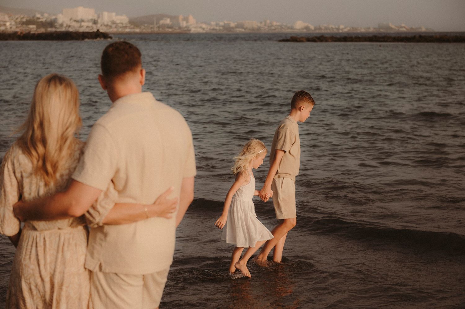 Parents in embrace watching children walk by ocean at sunset, Playa del Duque