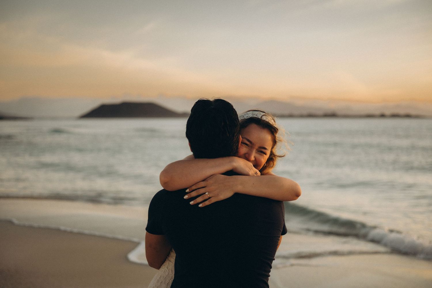Couple embracing on a beach at sunset with ocean waves and mountain silhouette in the background.