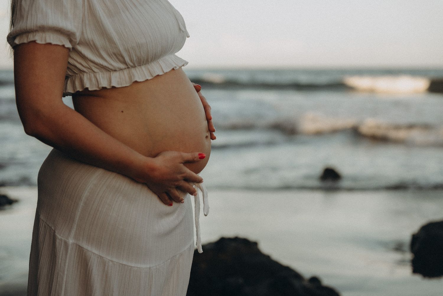 Pregnant woman in white dress cradling baby bump on rocky ocean beach at sunset.
