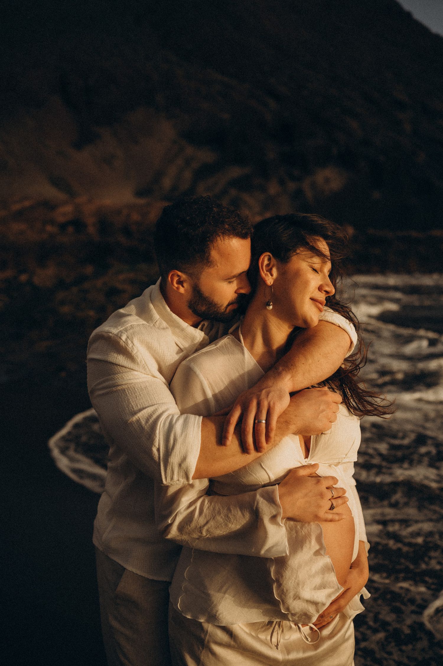 Couple embracing tenderly at golden hour on rocky coastline, man in white shirt holding woman close with ocean waves behind them.