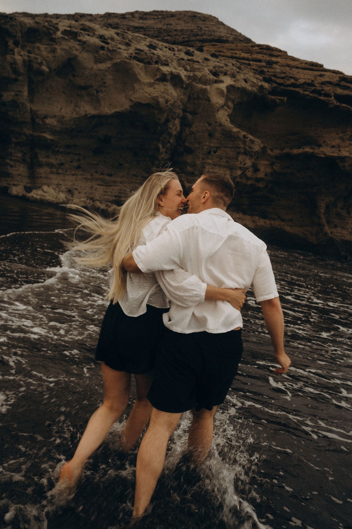 Couple in white shirts and black shorts embracing near ocean waves with rocky cliffs in background.
