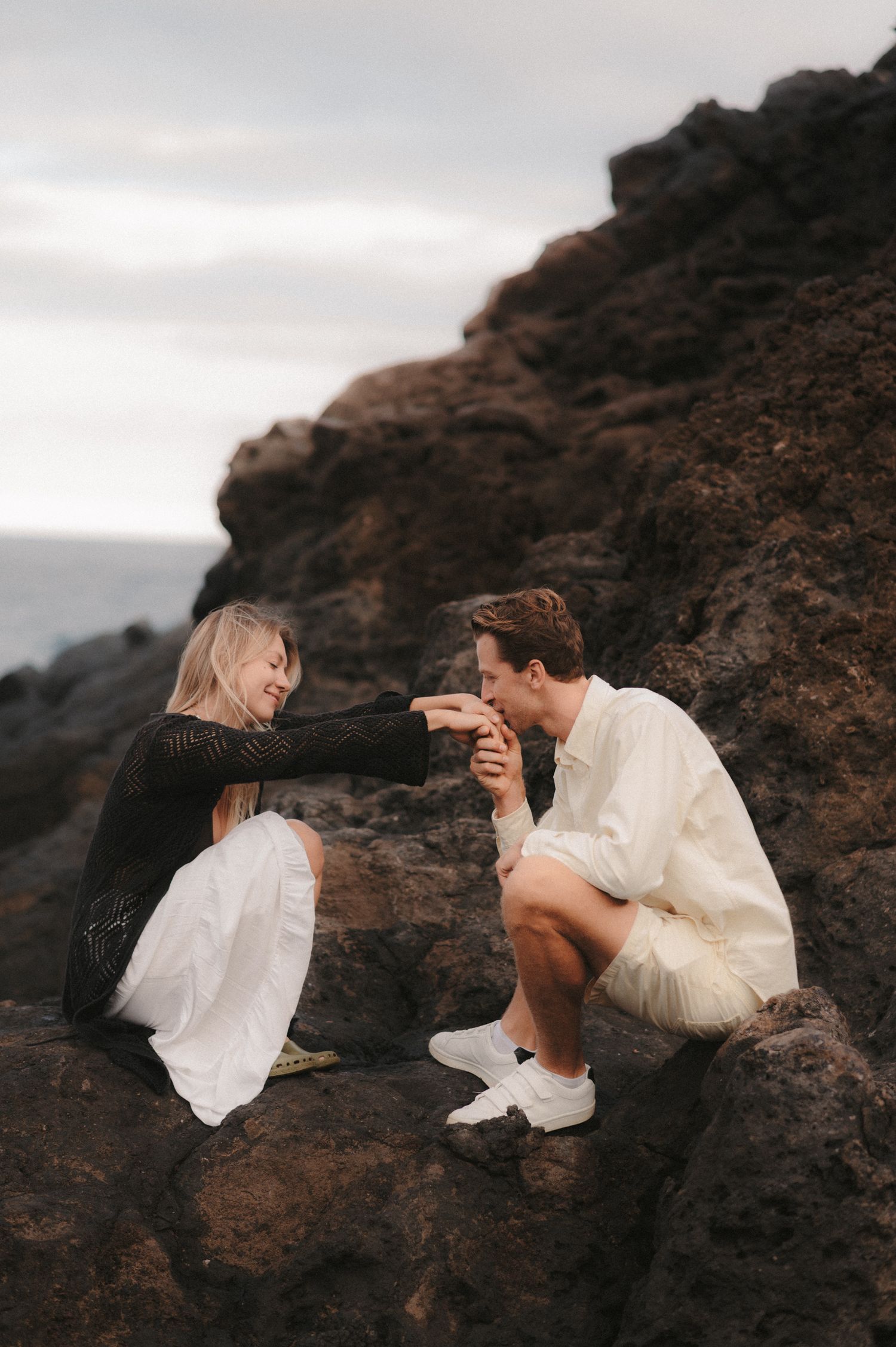 Loving couple sitting on rocks by the ocean in a secluded spot under Montaña Roja, he gently kissing her hand during a romantic photoshoot i