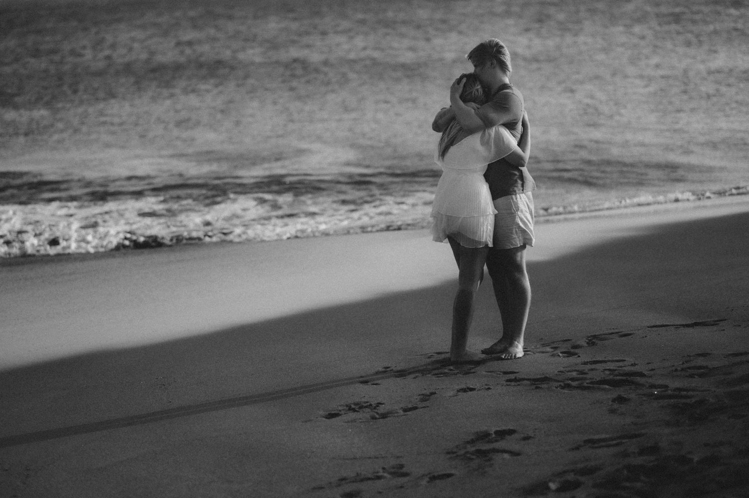 Young couple standing barefoot in the sand near the ocean, he holds her tightly in an embrace, with beautiful ocean and grand Playa Tejita i