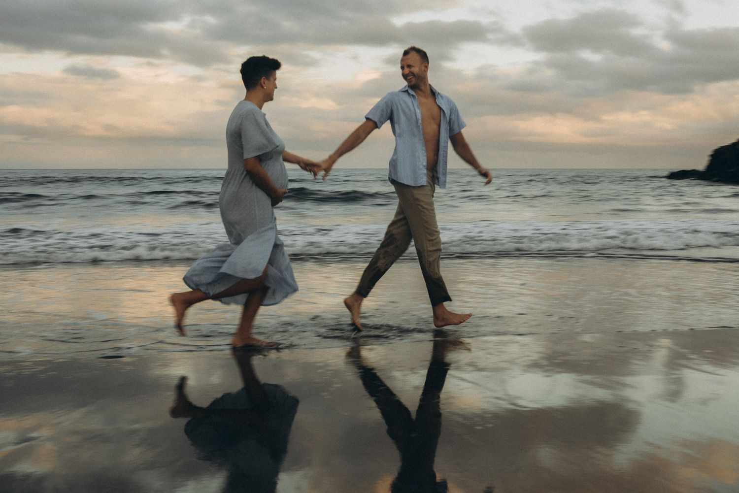 Couple holding hands running barefoot on a reflective beach at sunset with dramatic cloudy skies.