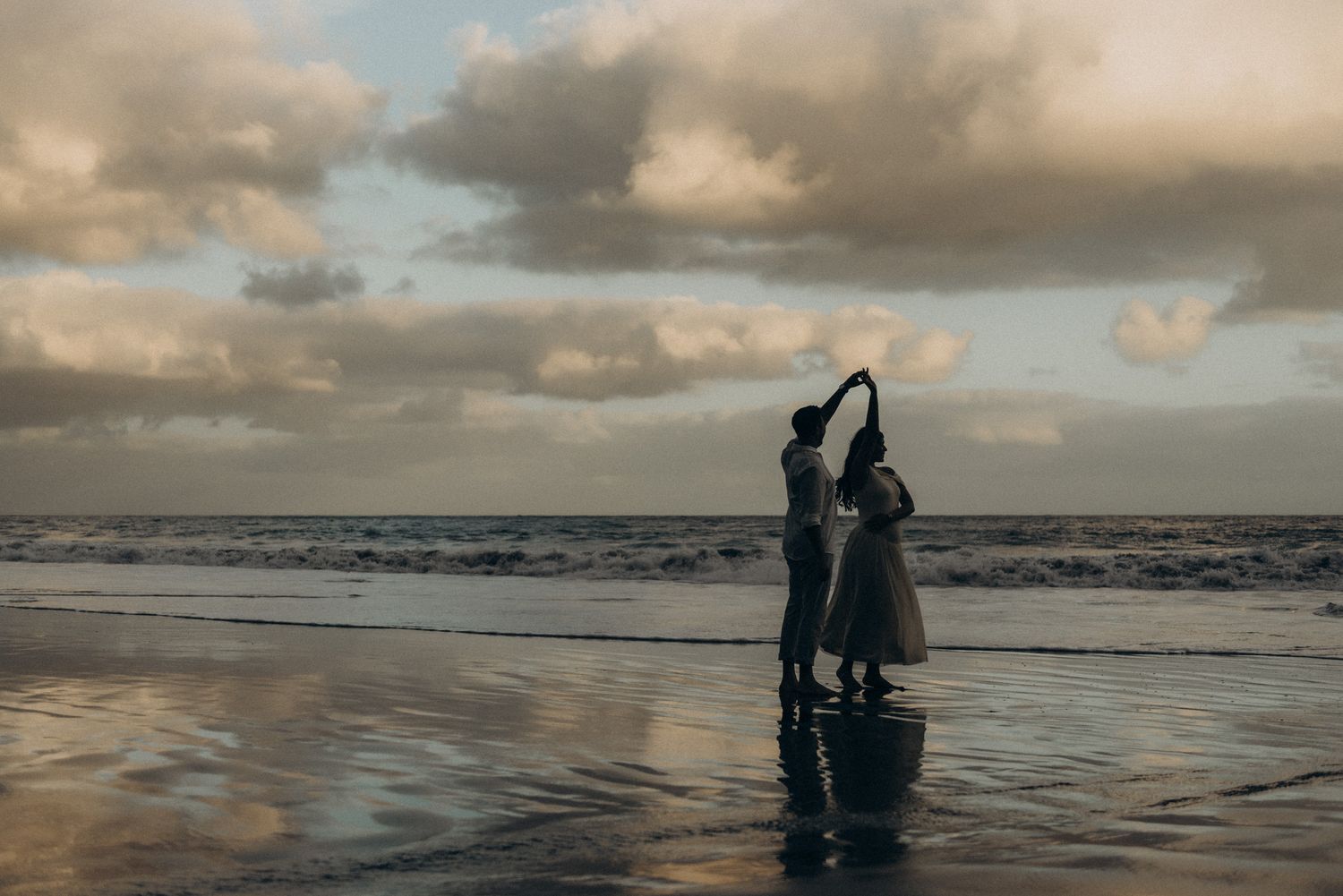 Couple dancing romantically on a reflective beach at sunset with dramatic cloudy sky and ocean waves.