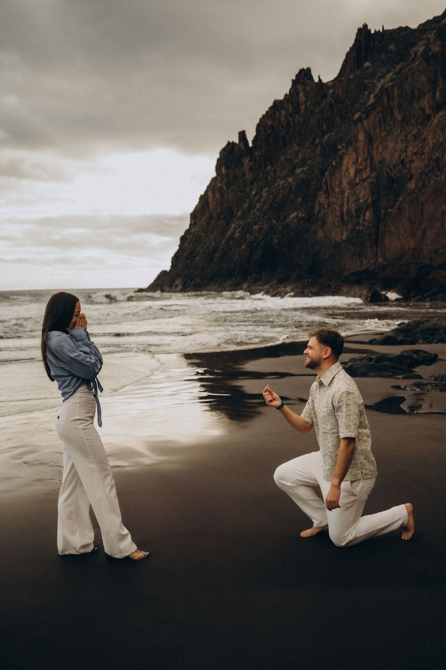 Romantic surprise proposal in Tenerife on a dramatic black sand beach with cliffs and ocean waves.