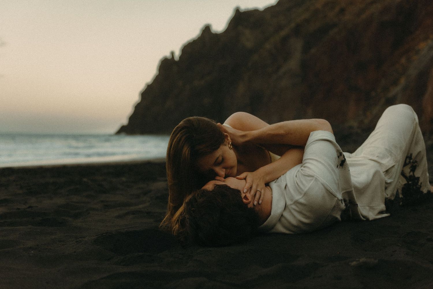 Romantic couple sharing an intimate moment lying on a dark sand beach at dusk with ocean cliffs in background.
